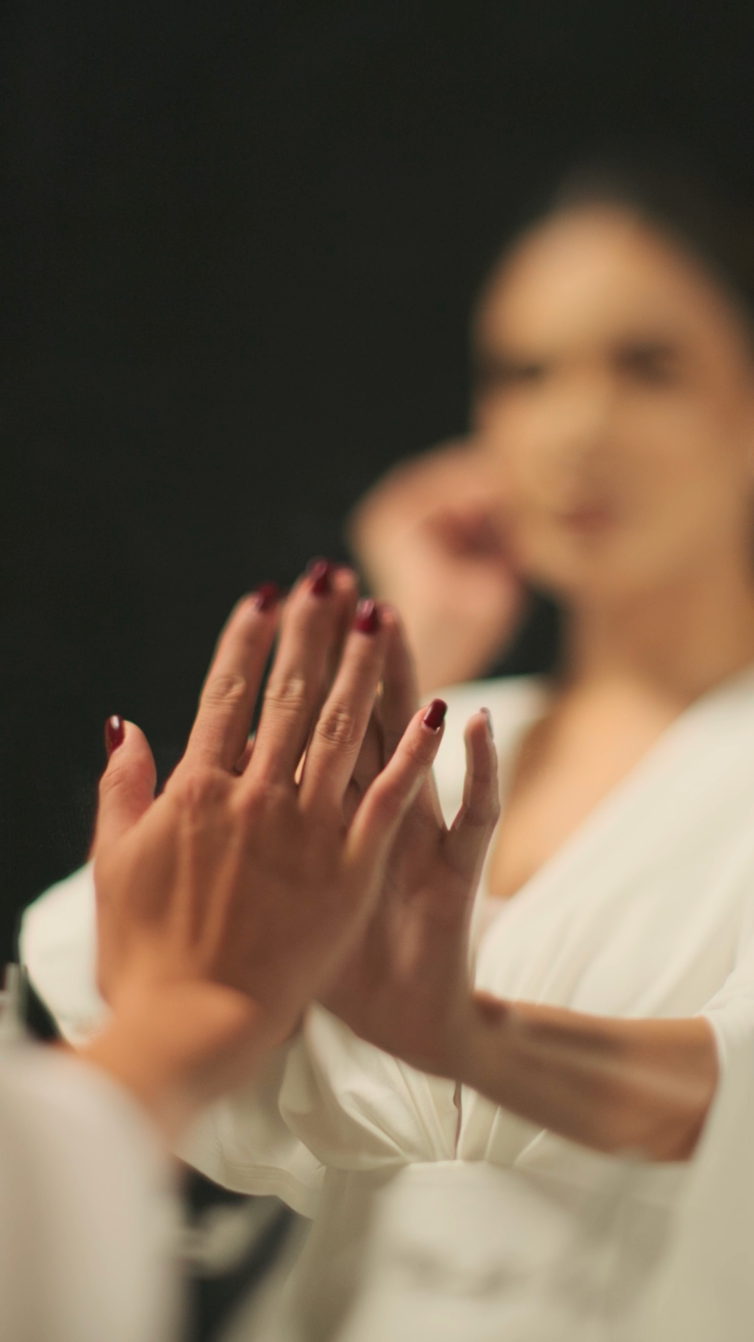 Close-up of womans hands with dark red manicured nails pressed together wearing a white robe in soft focus.
