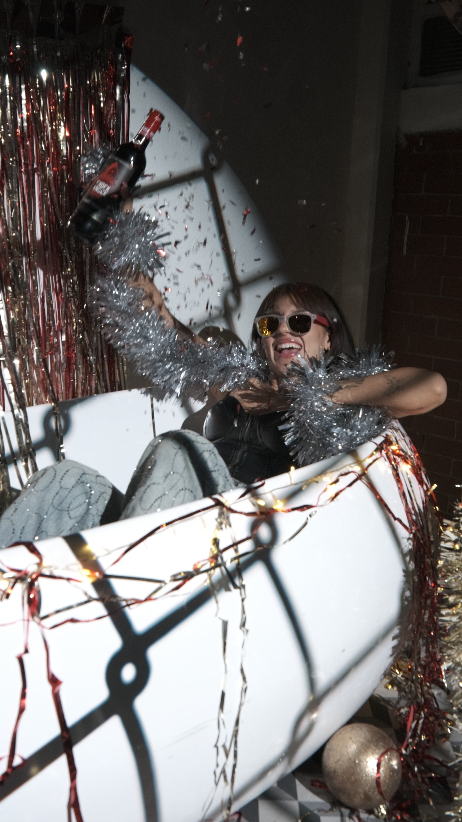 Smiling woman in sunglasses holding a bottle amid falling confetti and silver tinsel at a New Year's celebration.