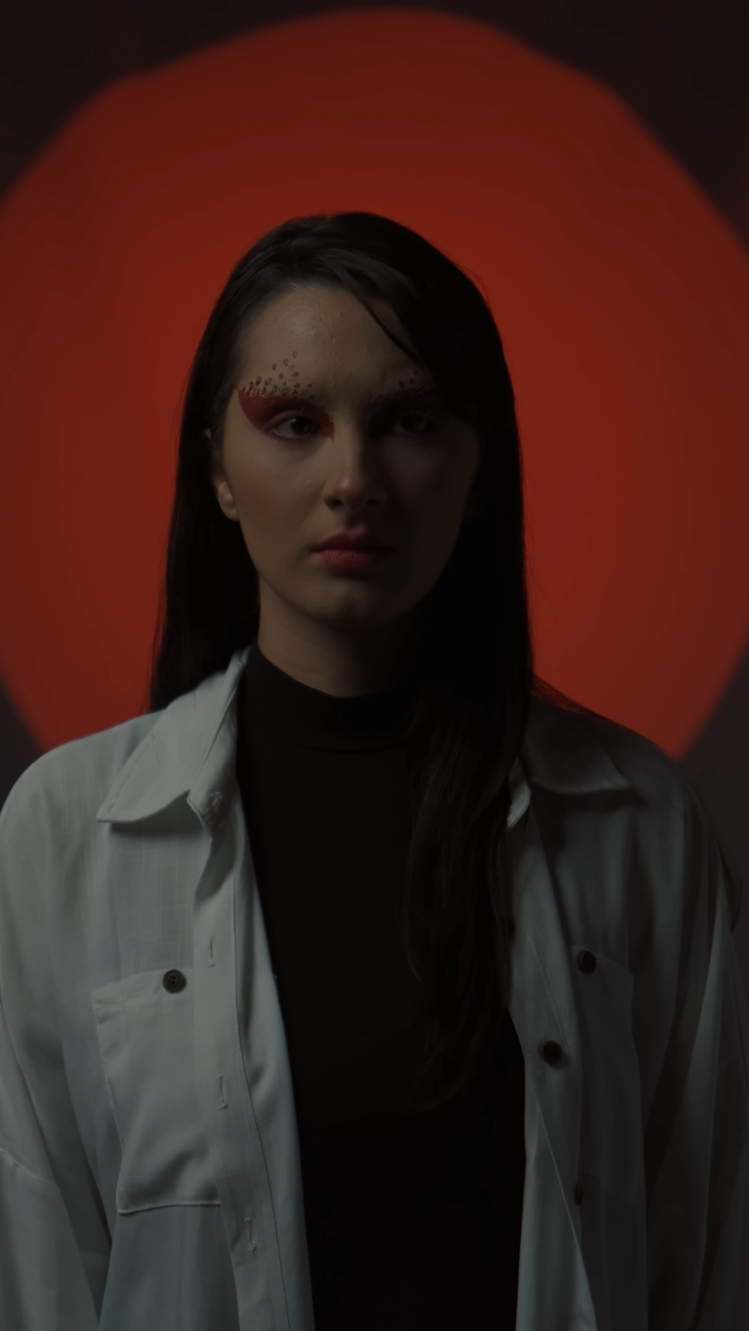 Young woman with dramatic red eye makeup in a white jacket poses before a red circular backdrop.
