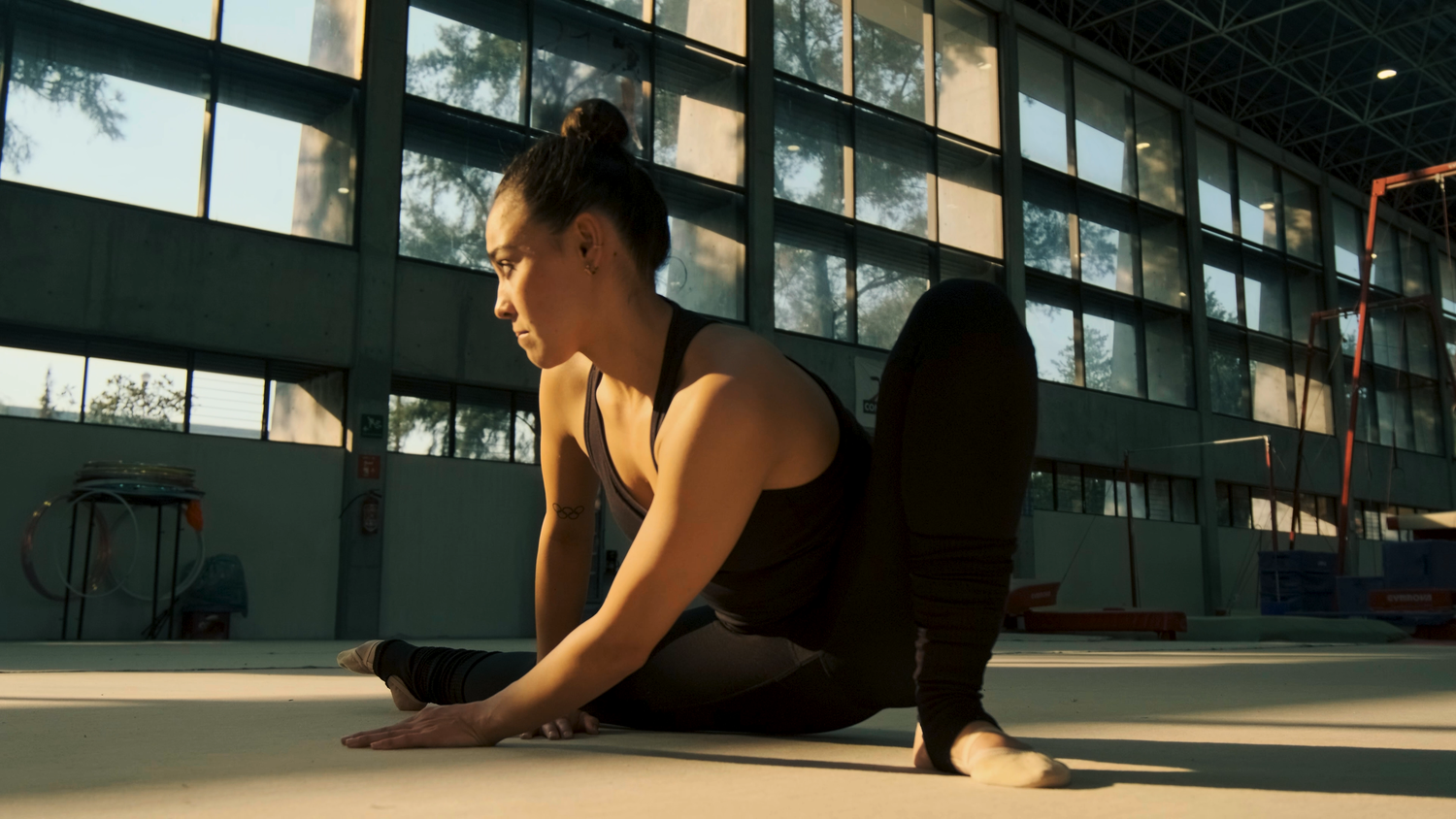 A female gymnast stretches gracefully on the floor inside a sunlit indoor gymnasium.