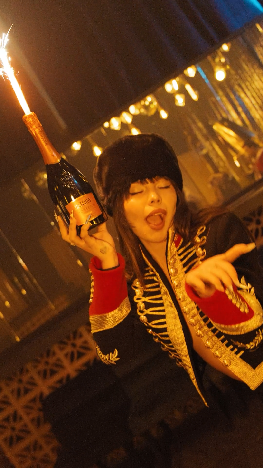 Person in decorative military jacket poses with a sparkling champagne bottle with sparkler at night event.