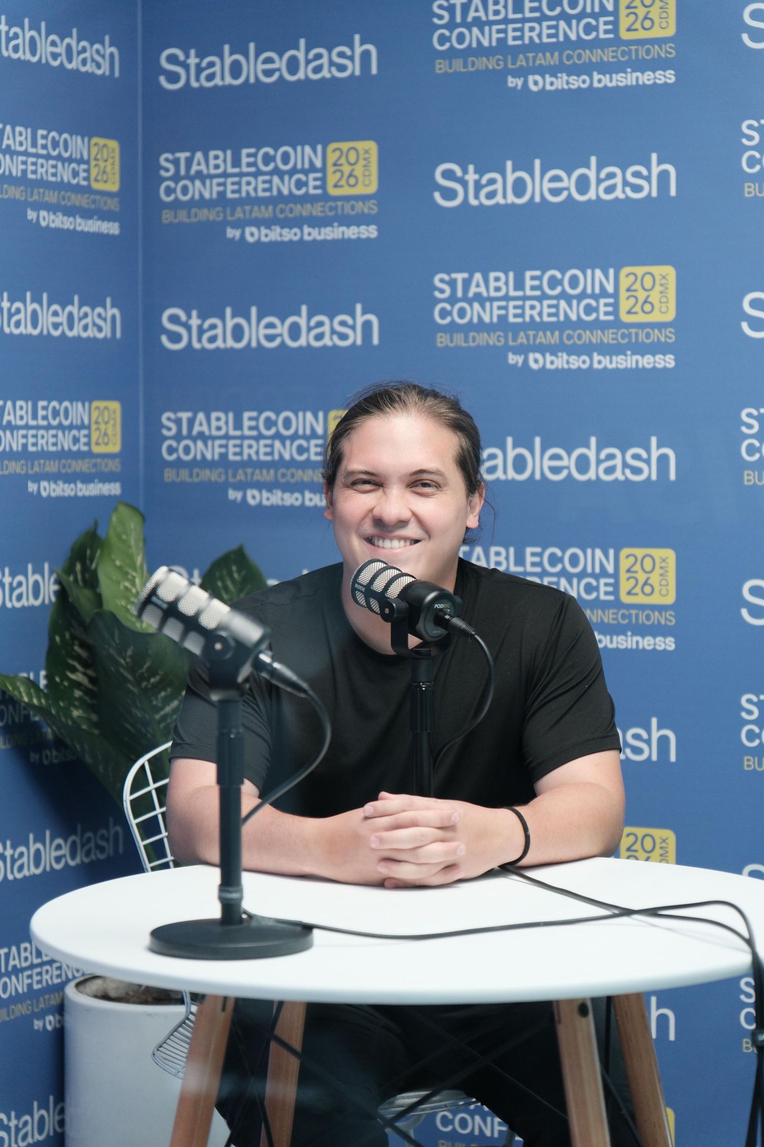 A smiling man sits at a podcast microphone in front of Stabledash Stablecoin Conference branded banners.