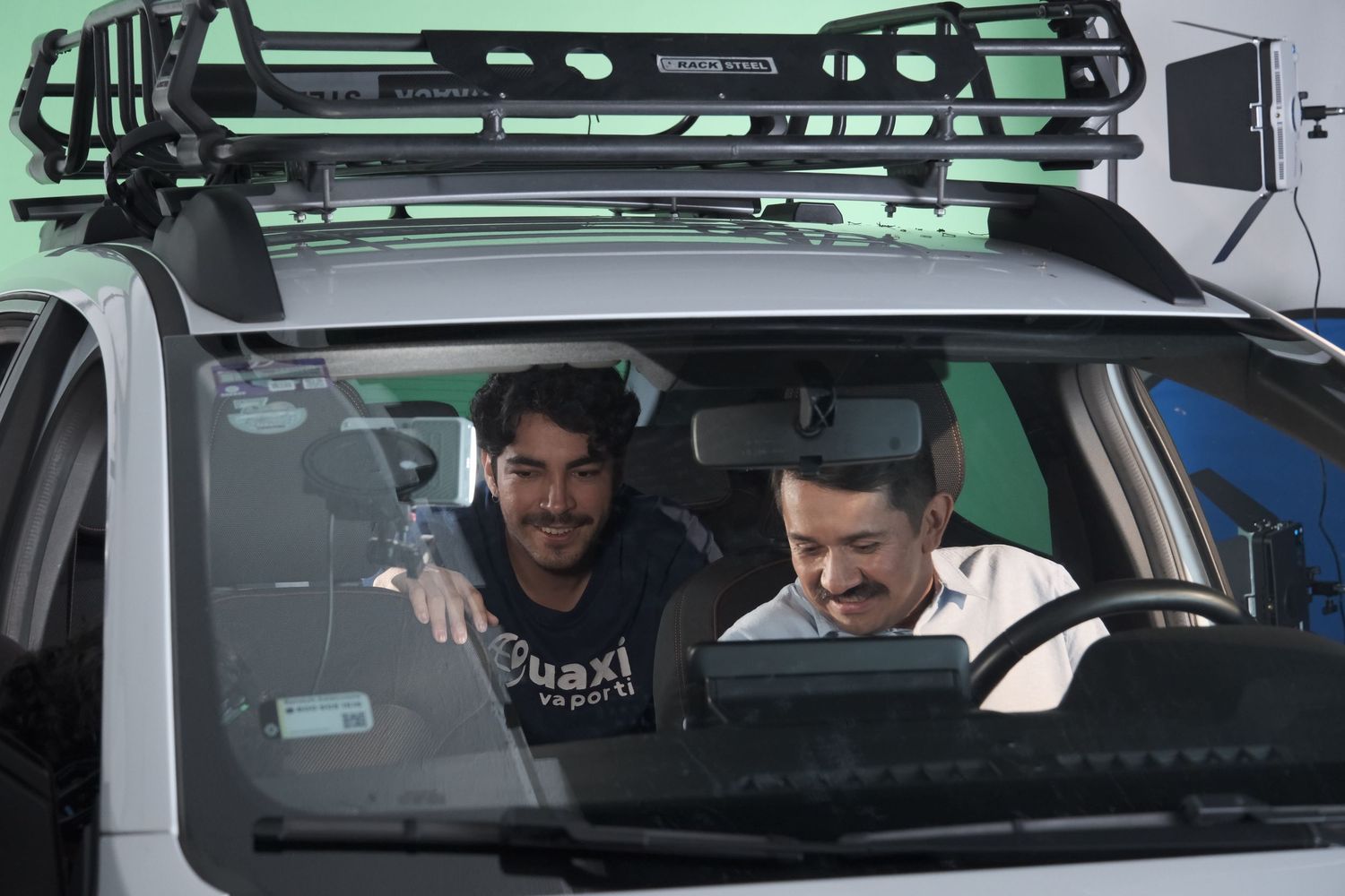 Two men reviewing content inside a vehicle equipped with roof rack and camera equipment on a film set.
