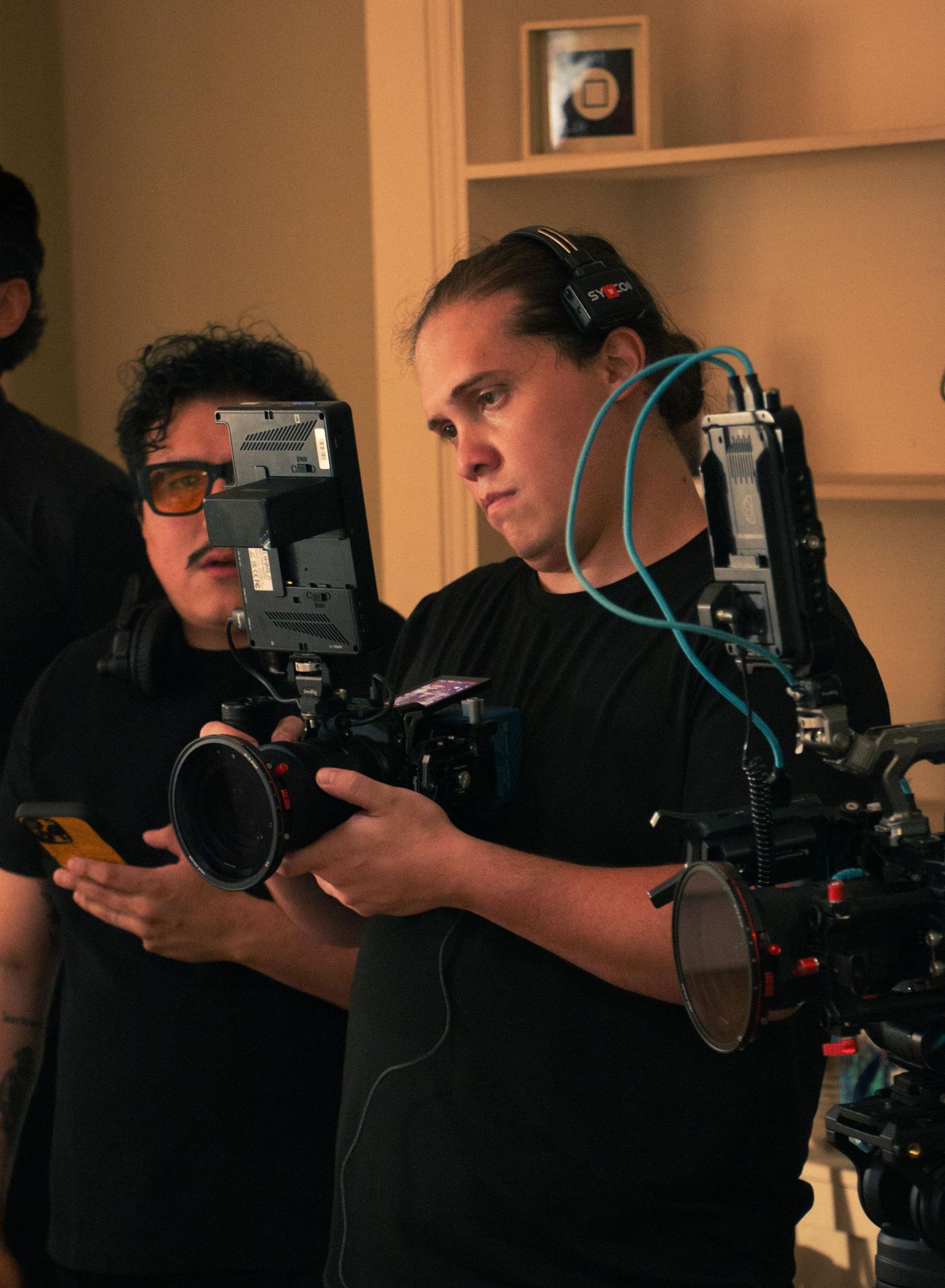Two film crew members operating a professional camera on set, one wearing headphones and a camera rig.