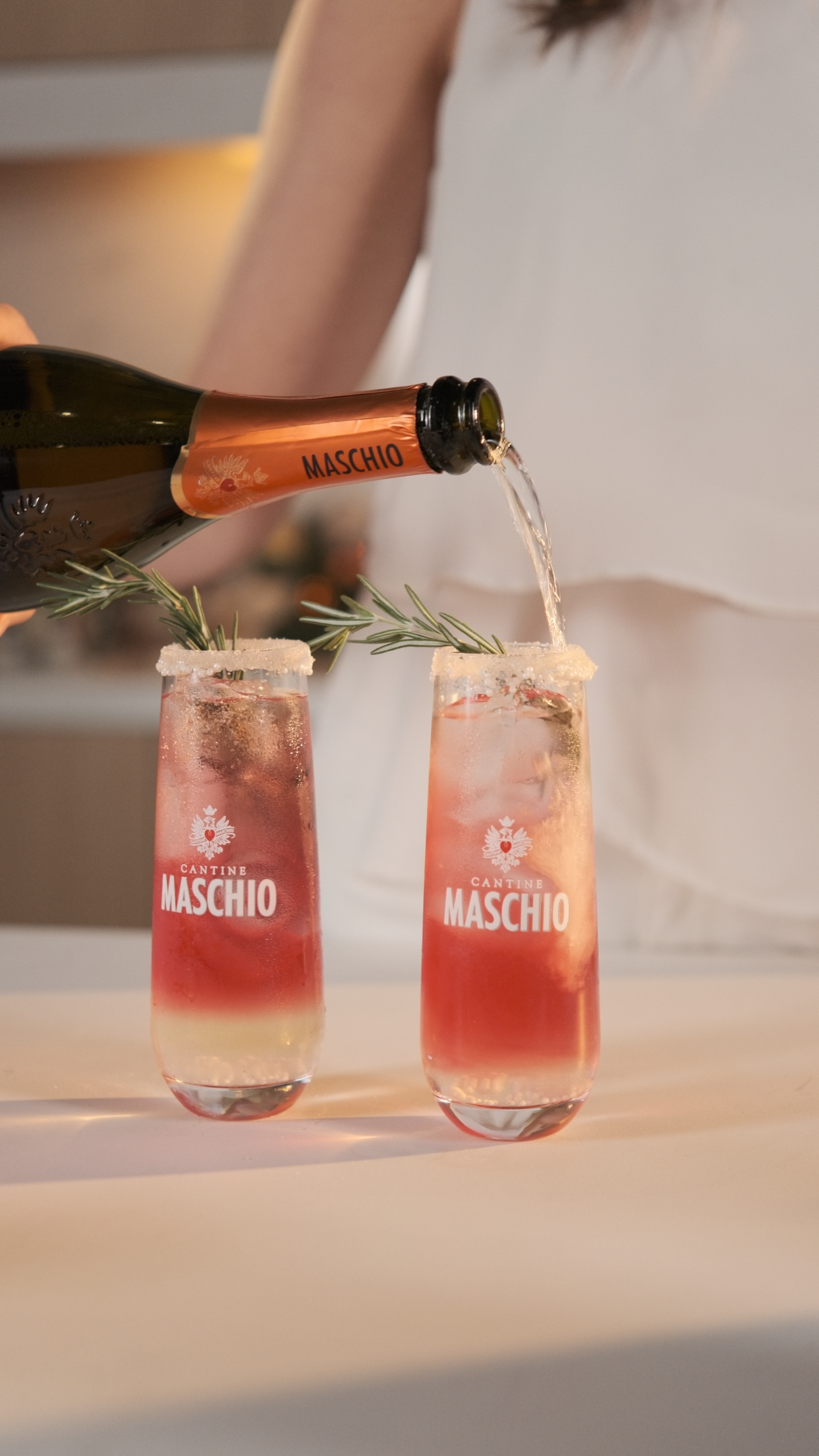 Maschio Prosecco being poured into two branded cocktail glasses with rosemary garnish.