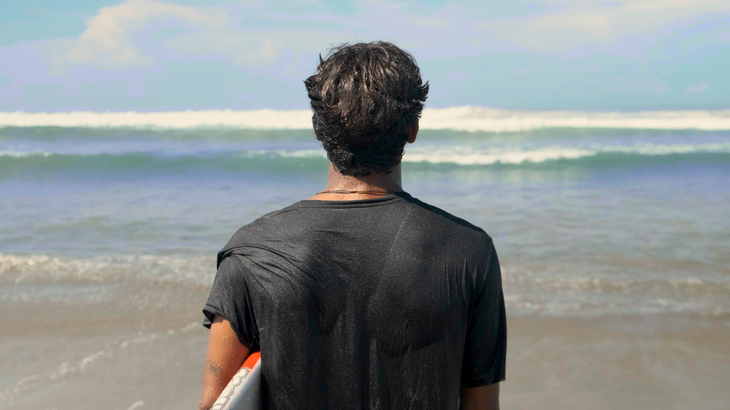 Man in black t-shirt standing on beach holding surfboard looking out at ocean waves.