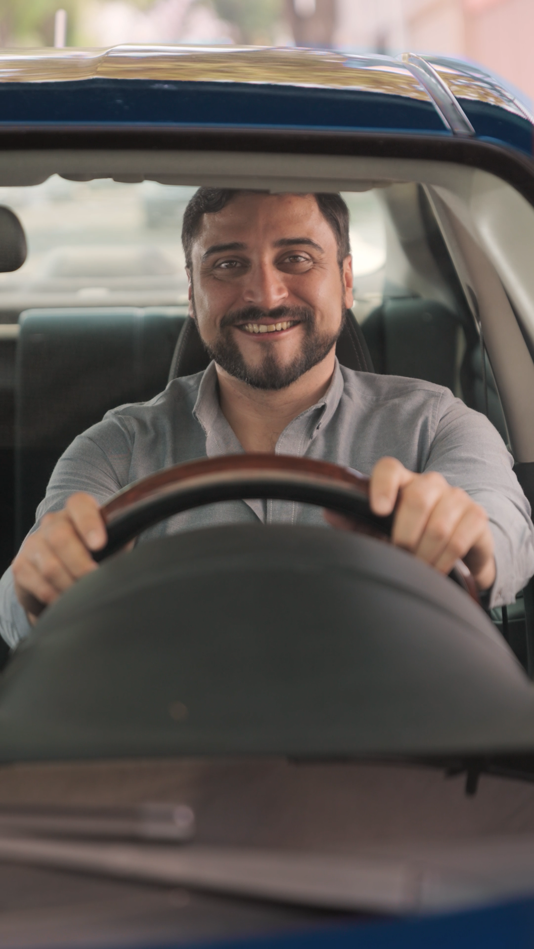 Smiling bearded man in gray shirt sitting behind steering wheel of blue car.