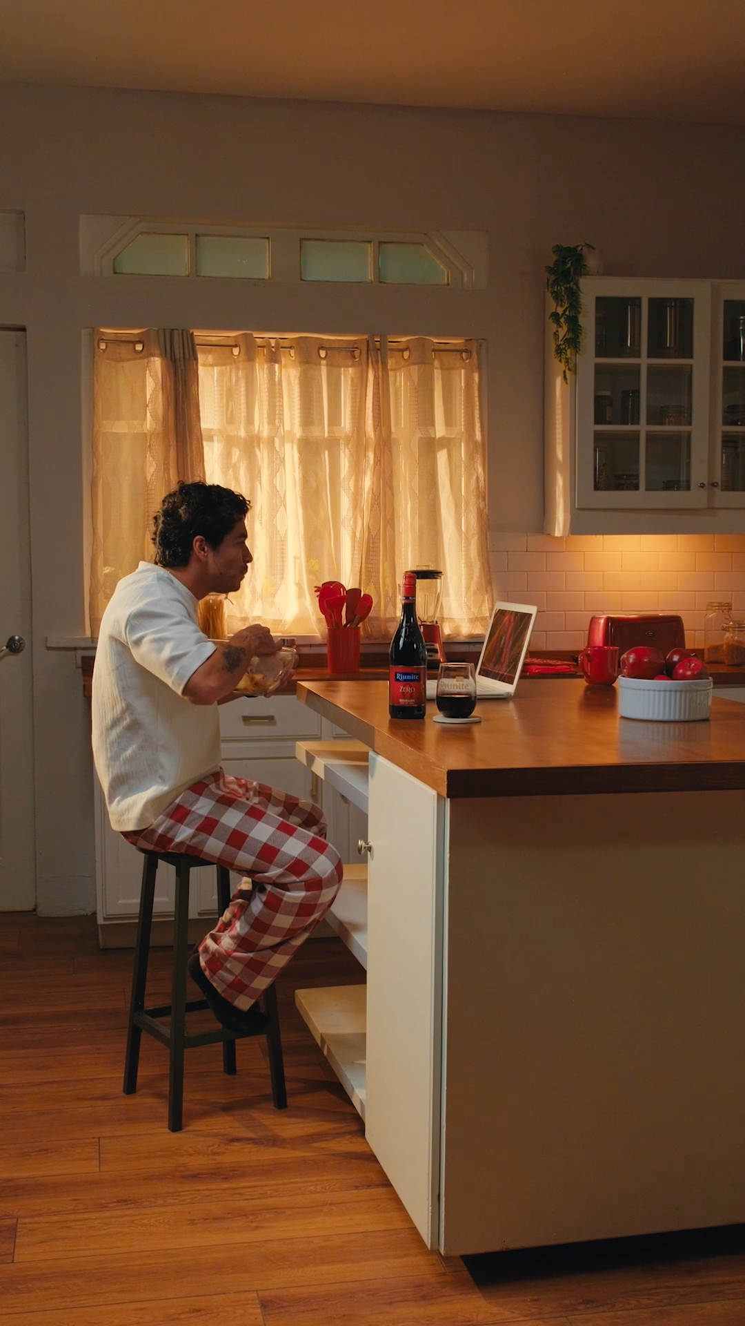 Man in plaid pajama pants sitting at kitchen counter with wine bottle and laptop.