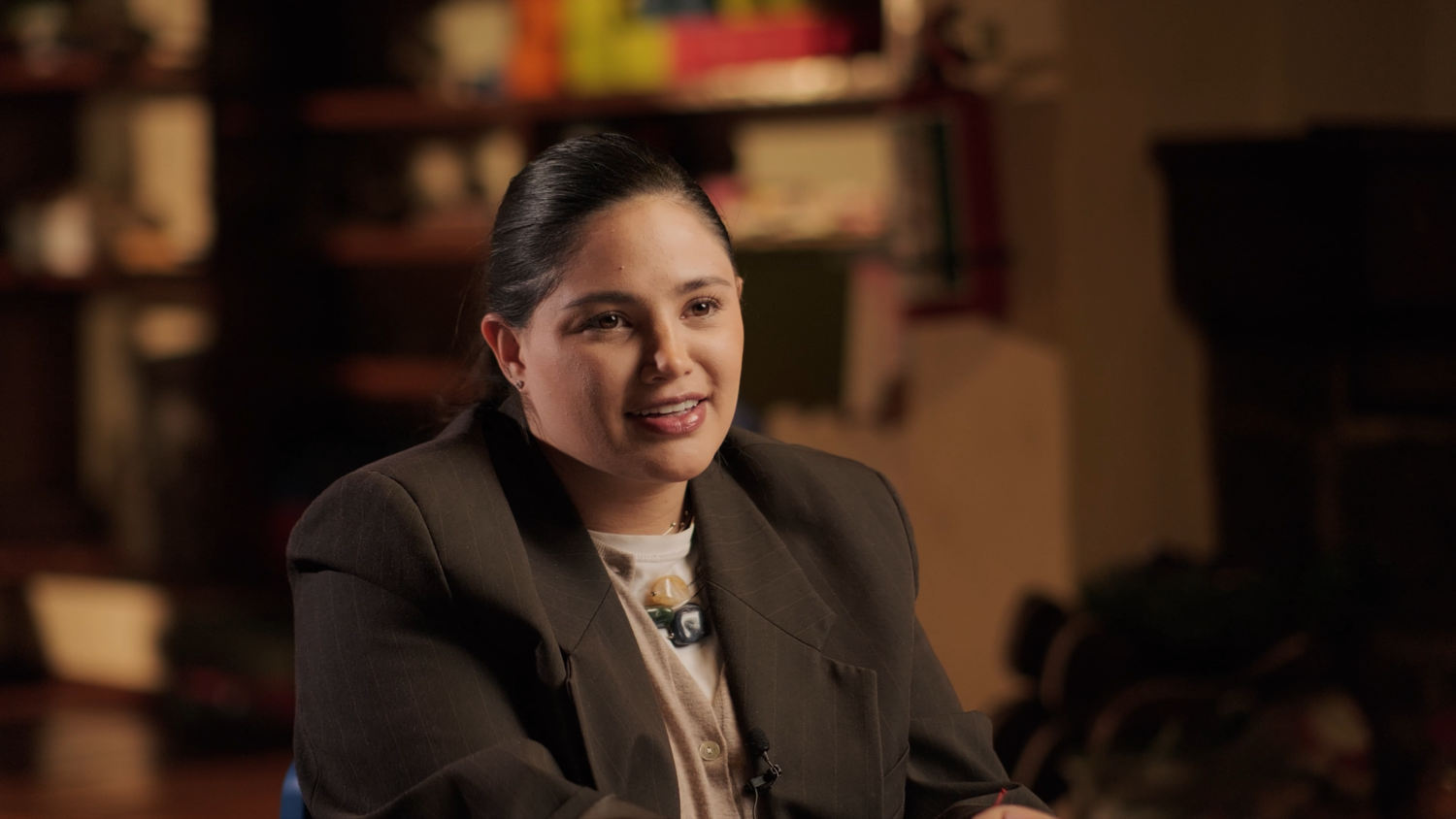 Smiling woman in a brown blazer sitting in a warmly lit room with bookshelves behind her.