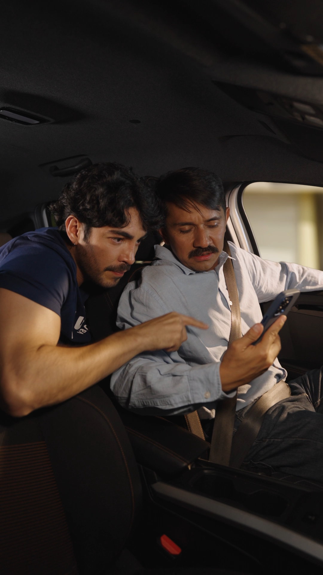 Two men inside a car at night, one pointing at a smartphone screen held by the other.