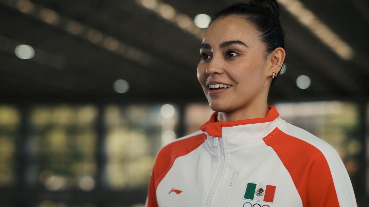 Young woman smiling in a Mexican Olympic team white and red athletic jacket indoors.