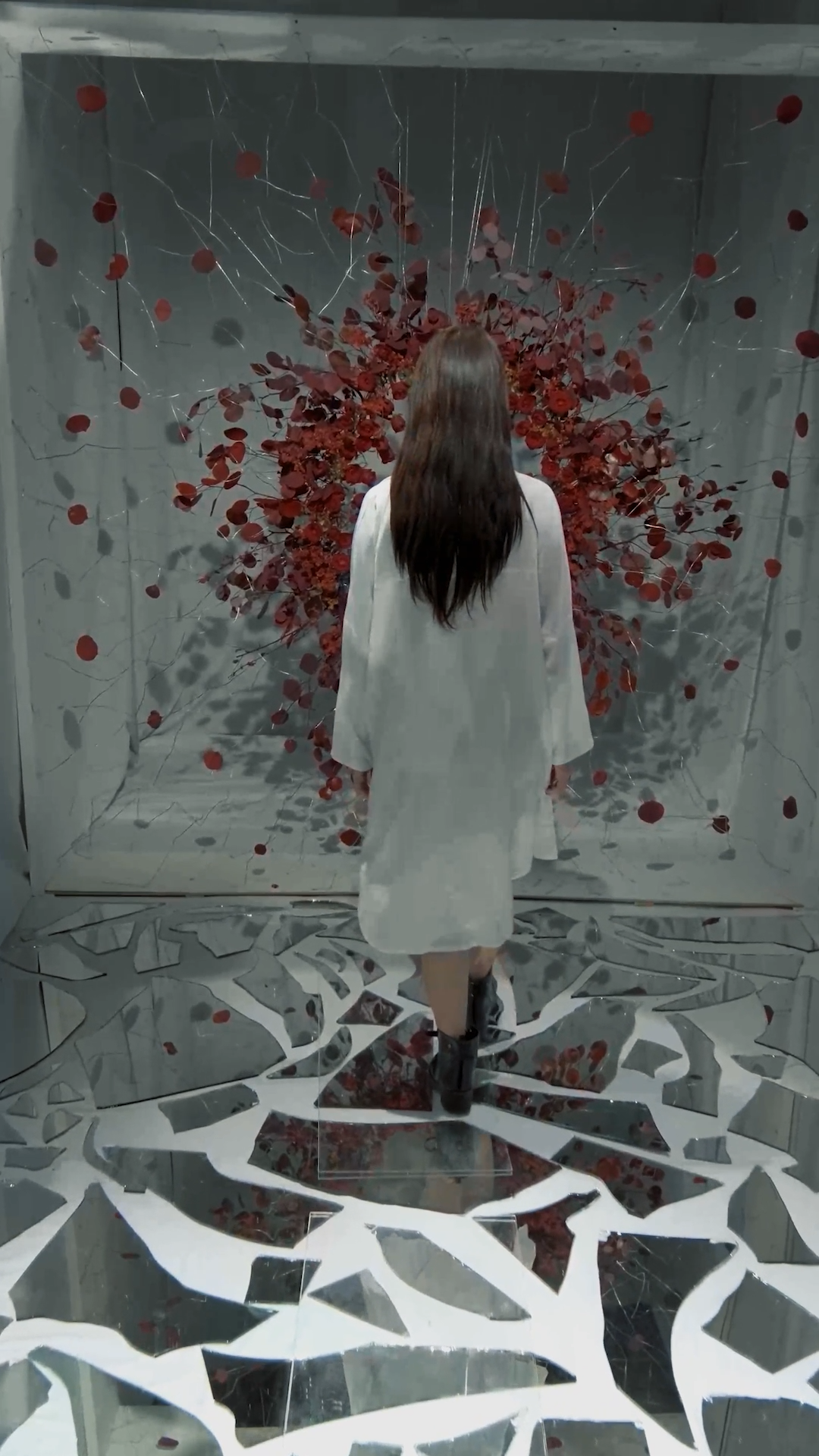 Woman in white dress stands before a dramatic wall installation of red roses with shattered mirror floor pieces.
