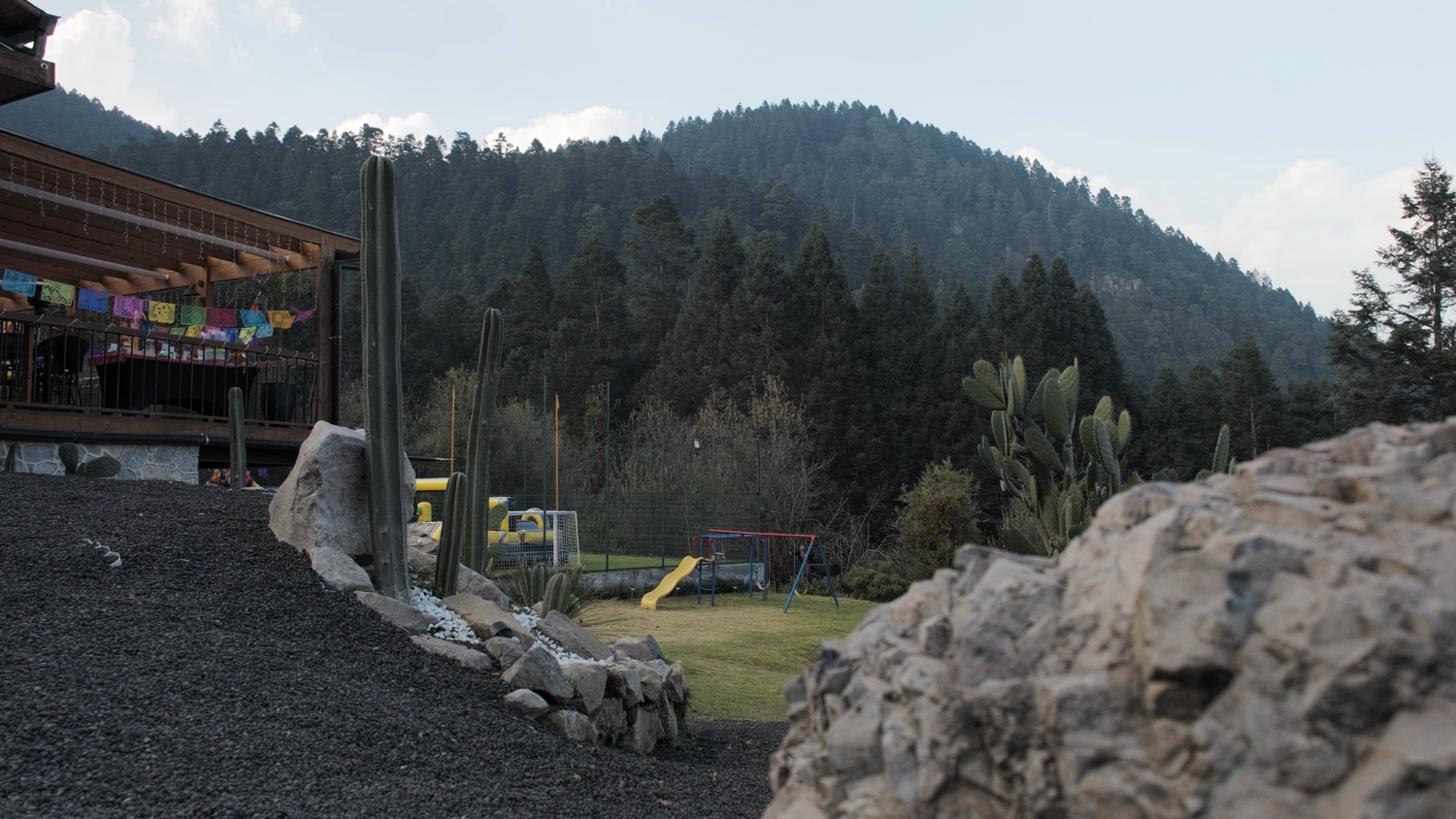 Scenic mountain landscape with pine forest, rocks, and colorful flags near a rustic building.