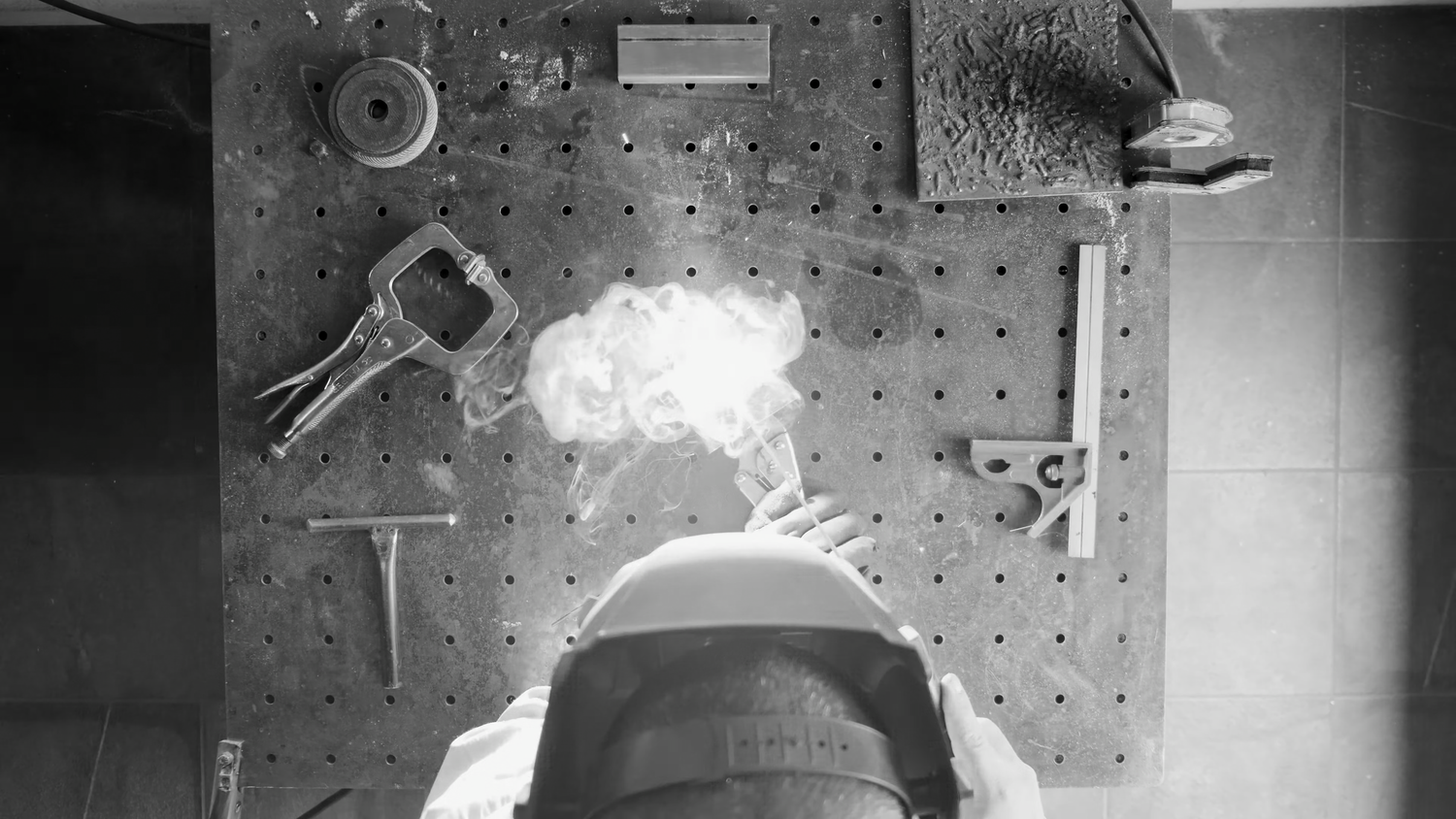 Overhead black and white photo of welder working on perforated metal surface with tools nearby.