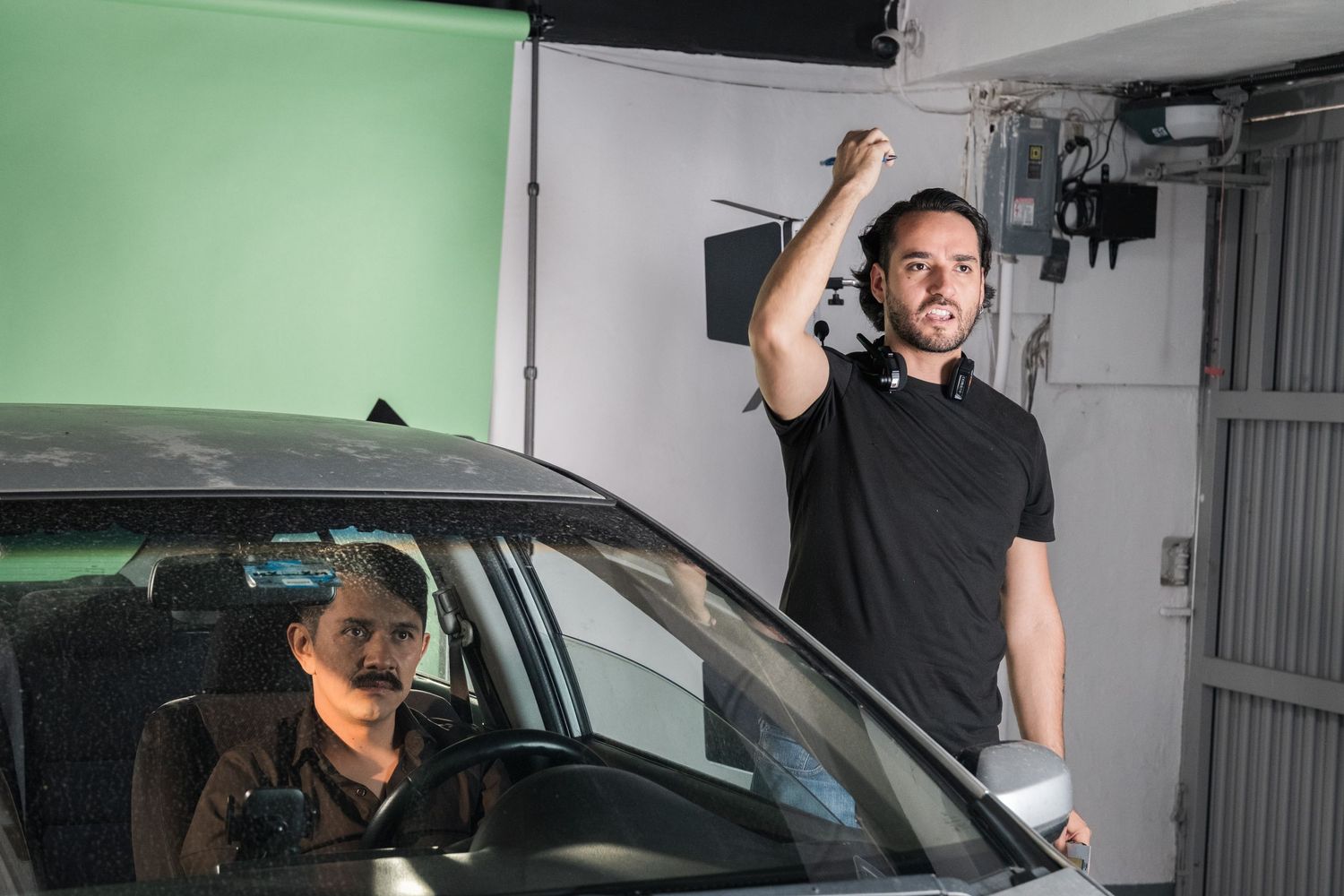 A film director stands beside a car on a green screen set, gesturing with his hand.