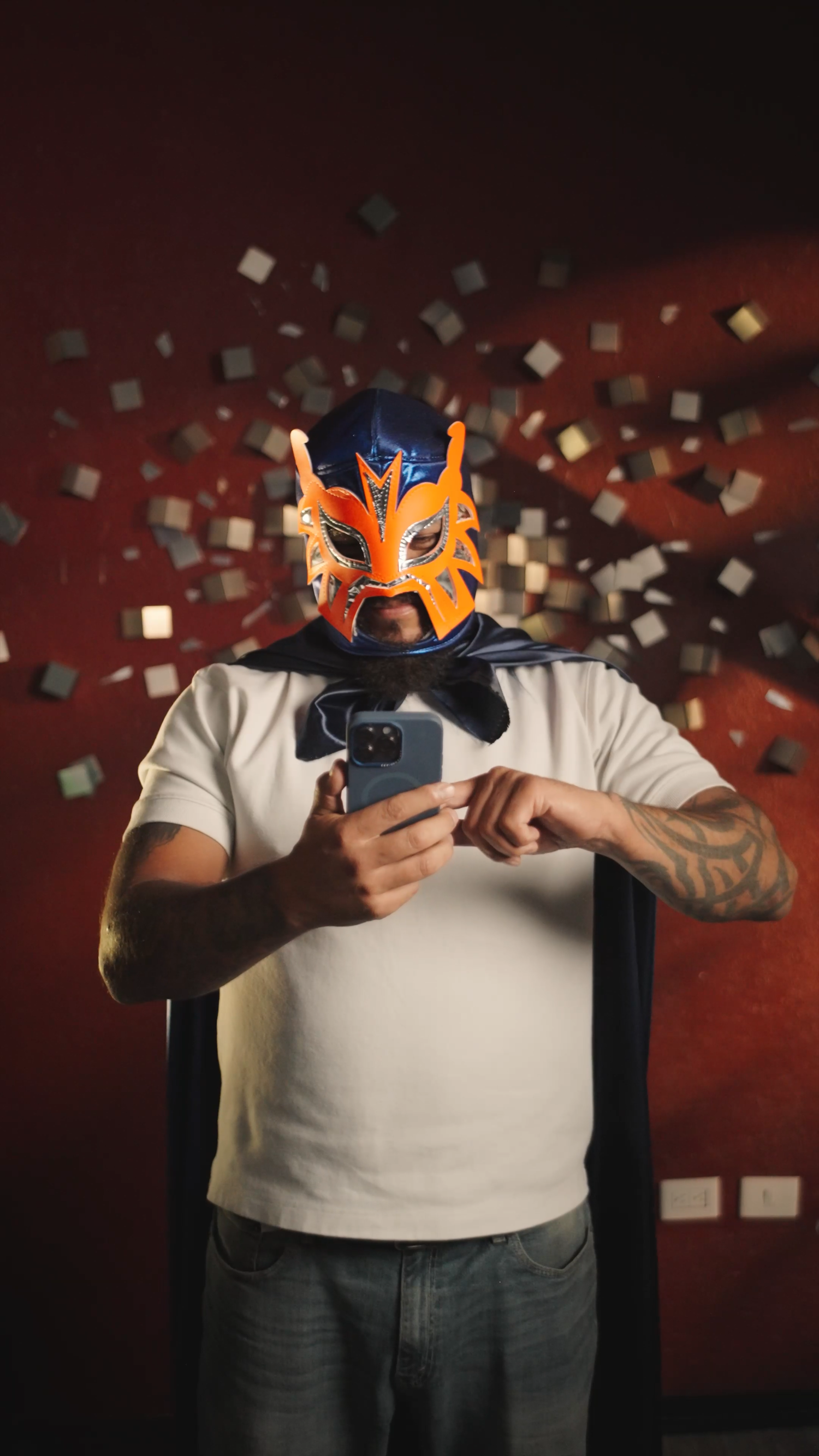 Tattooed man wearing colorful lucha libre wrestling mask using smartphone indoors.