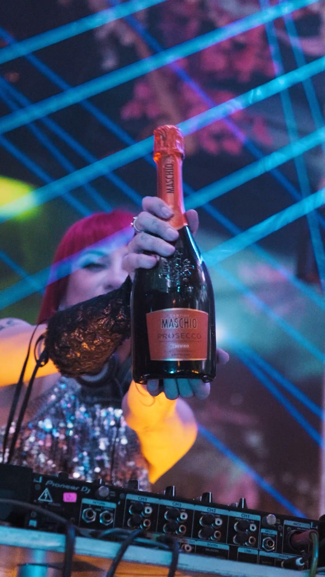 Female DJ in sequin outfit holds up a Maschio Prosecco bottle under colorful laser lights at a nightclub.