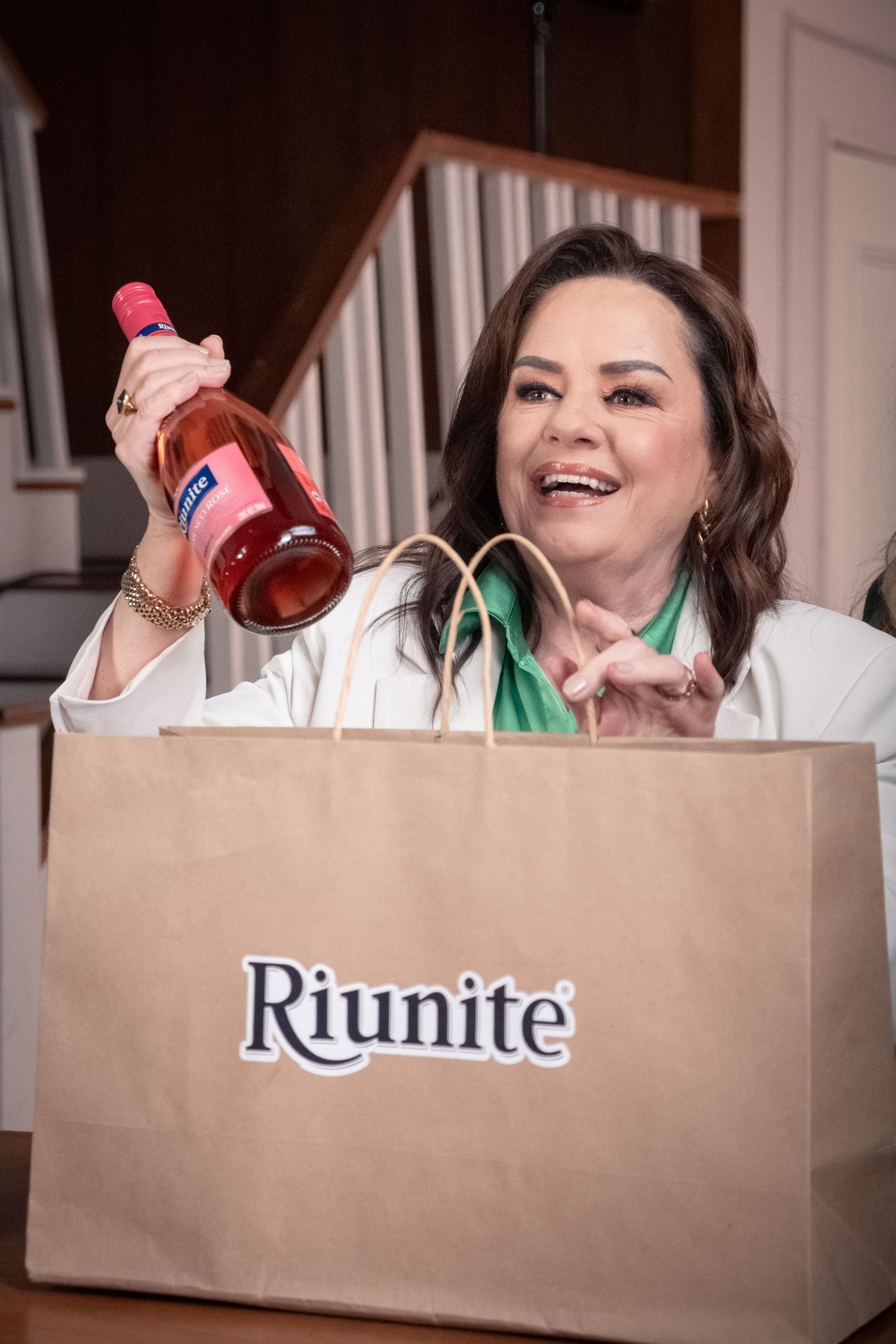 Smiling woman holding a Riunite rose wine bottle from a branded shopping bag near a staircase.