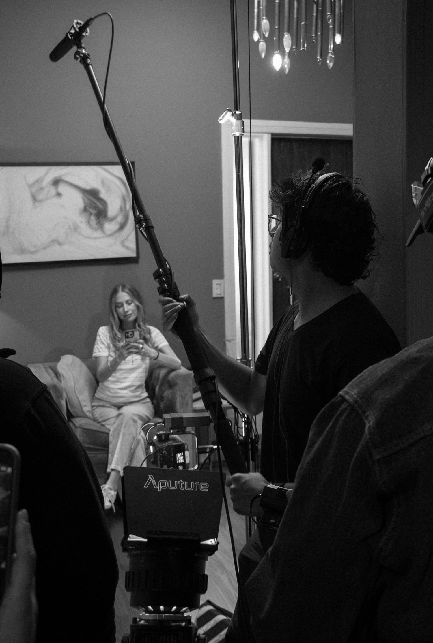 Black and white behind-the-scenes photo of crew filming actress on couch with boom mic and Aputure light.