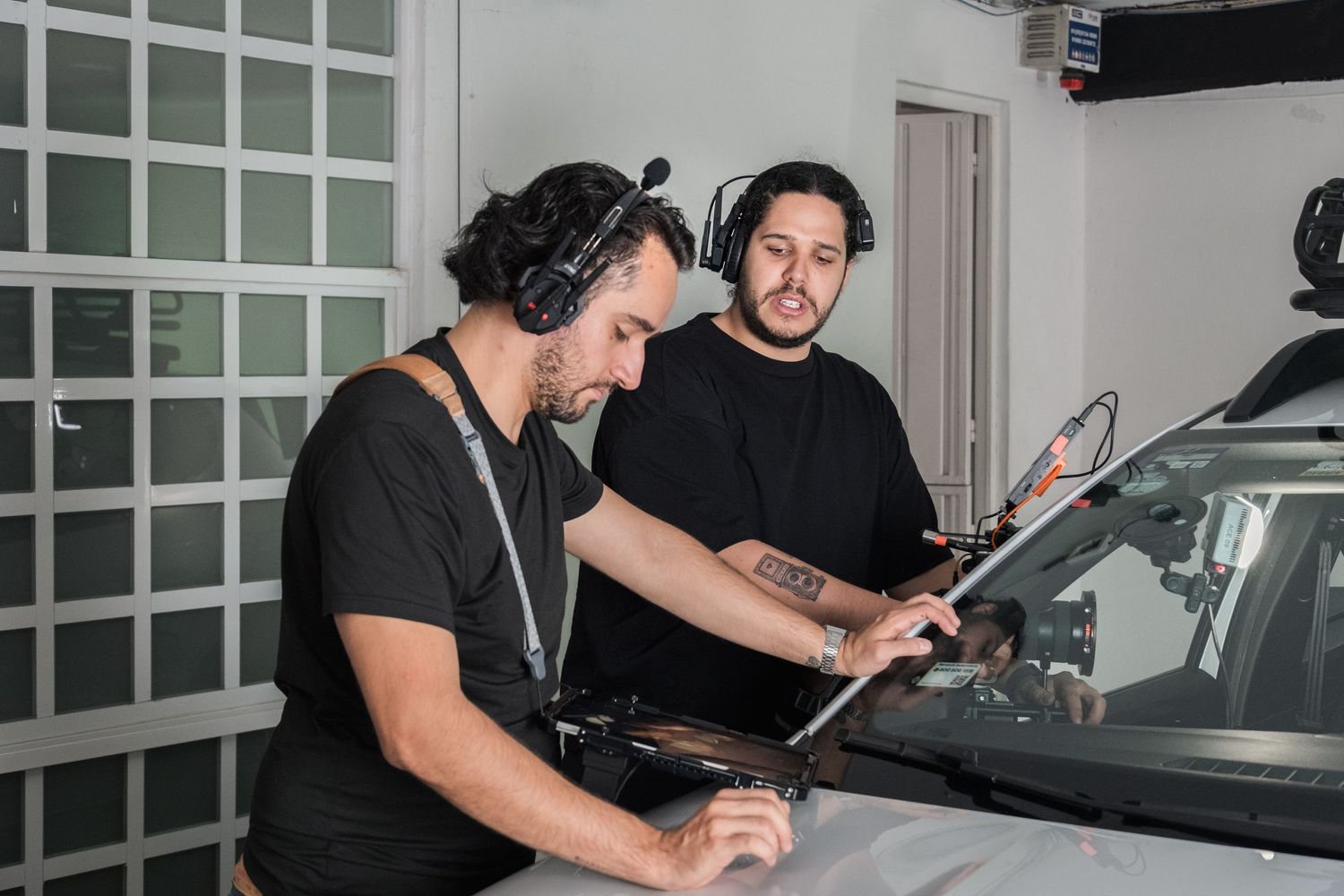 Two men in black shirts collaborate over a laptop on the hood of a car on a film set.