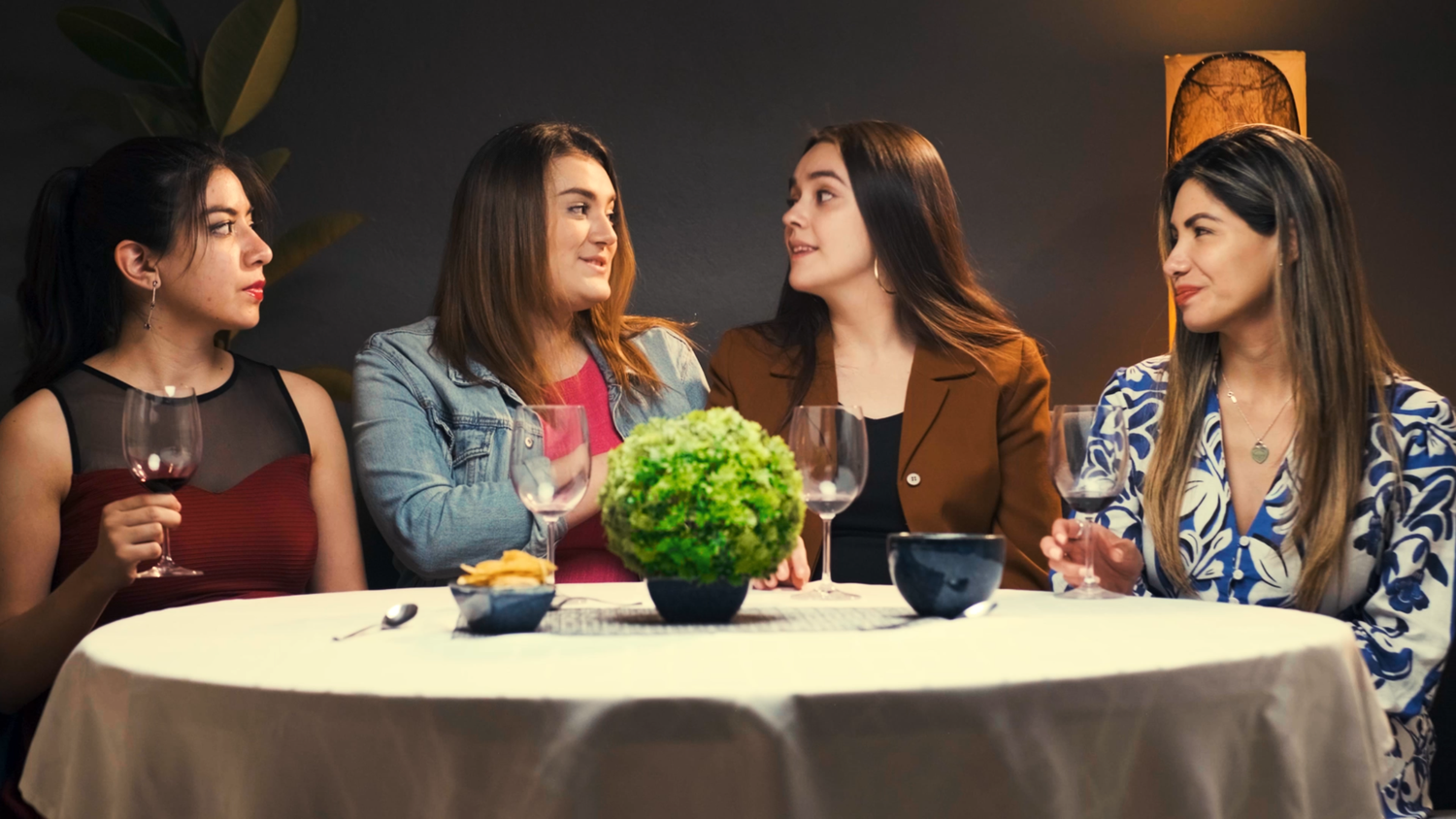 Four women sitting together at a dining table with wine glasses and a small plant centerpiece.
