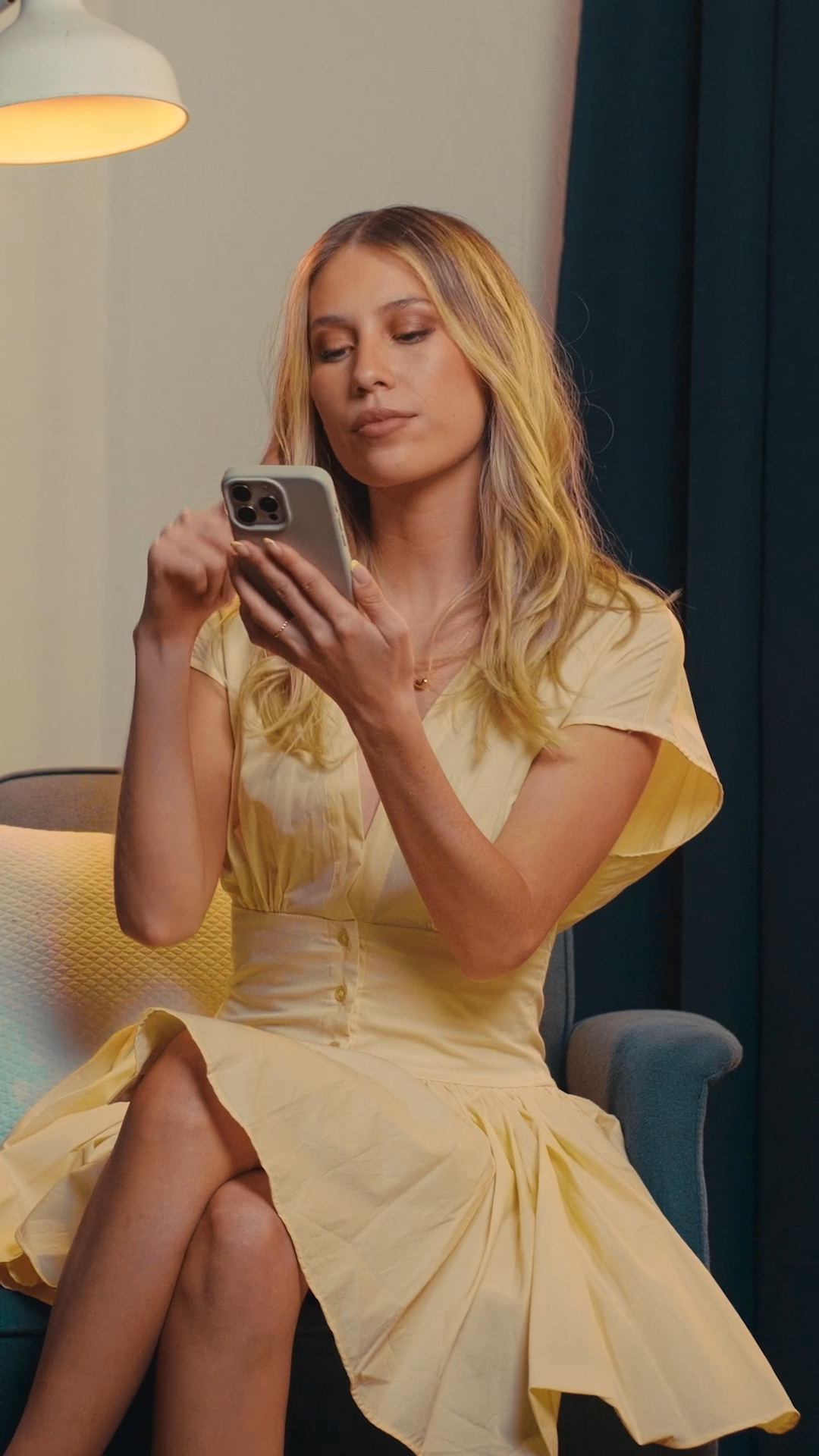 Young blonde woman in yellow dress using smartphone while seated in teal armchair indoors.