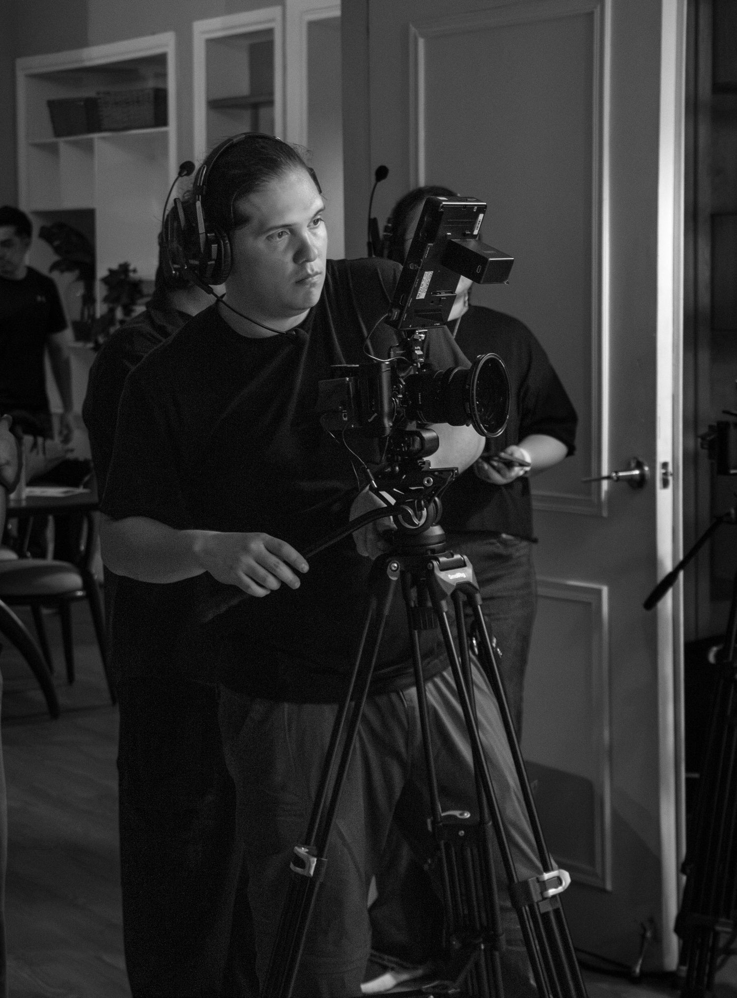 Black and white behind-the-scenes photo of a cameraman operating a professional film camera on a tripod.