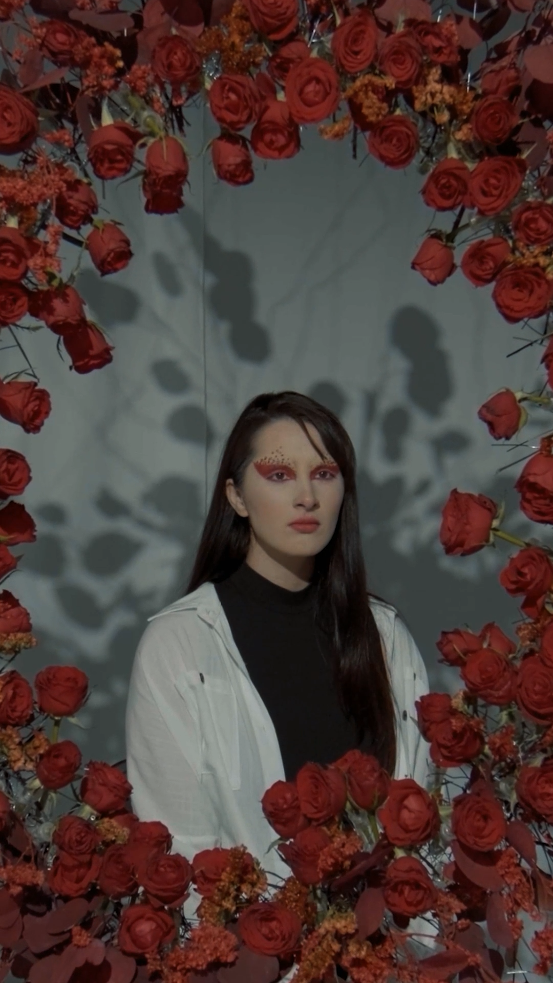 Woman with bold red eye makeup poses surrounded by a frame of vibrant red roses and orange flowers.