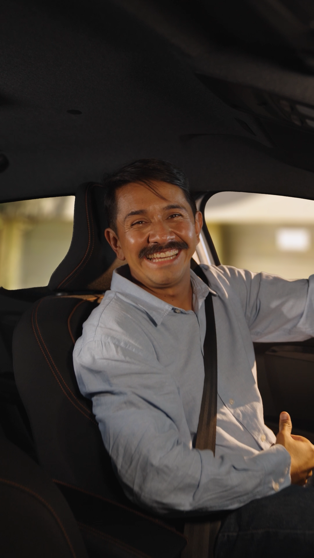 Smiling middle-aged man sitting in a car wearing a seatbelt and light blue shirt.