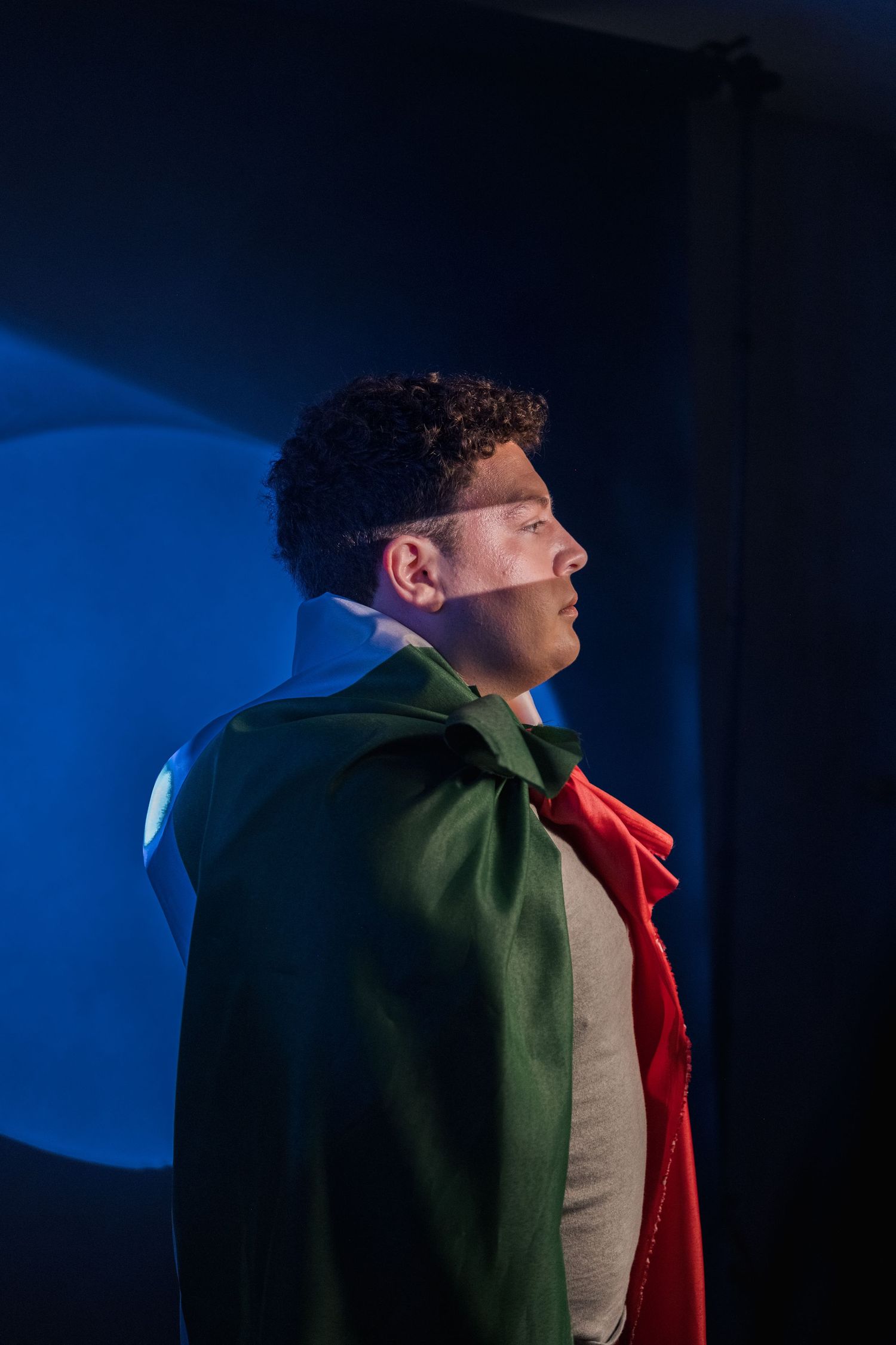 Man in green cape and red cravat poses in profile against dramatic blue lit background.
