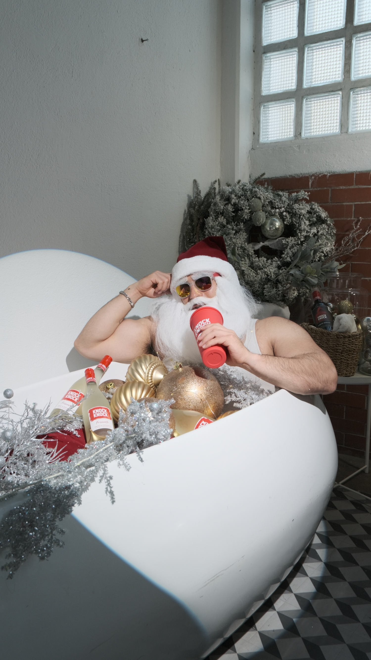 Muscular man in Santa costume relaxing in bathtub with red beverage bottles and gold Christmas ornaments.