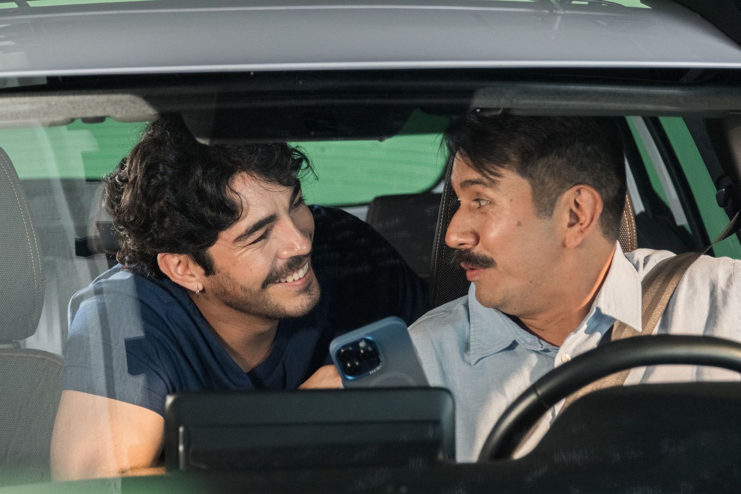 Two men with mustaches share a conversation inside a car in a film production scene.