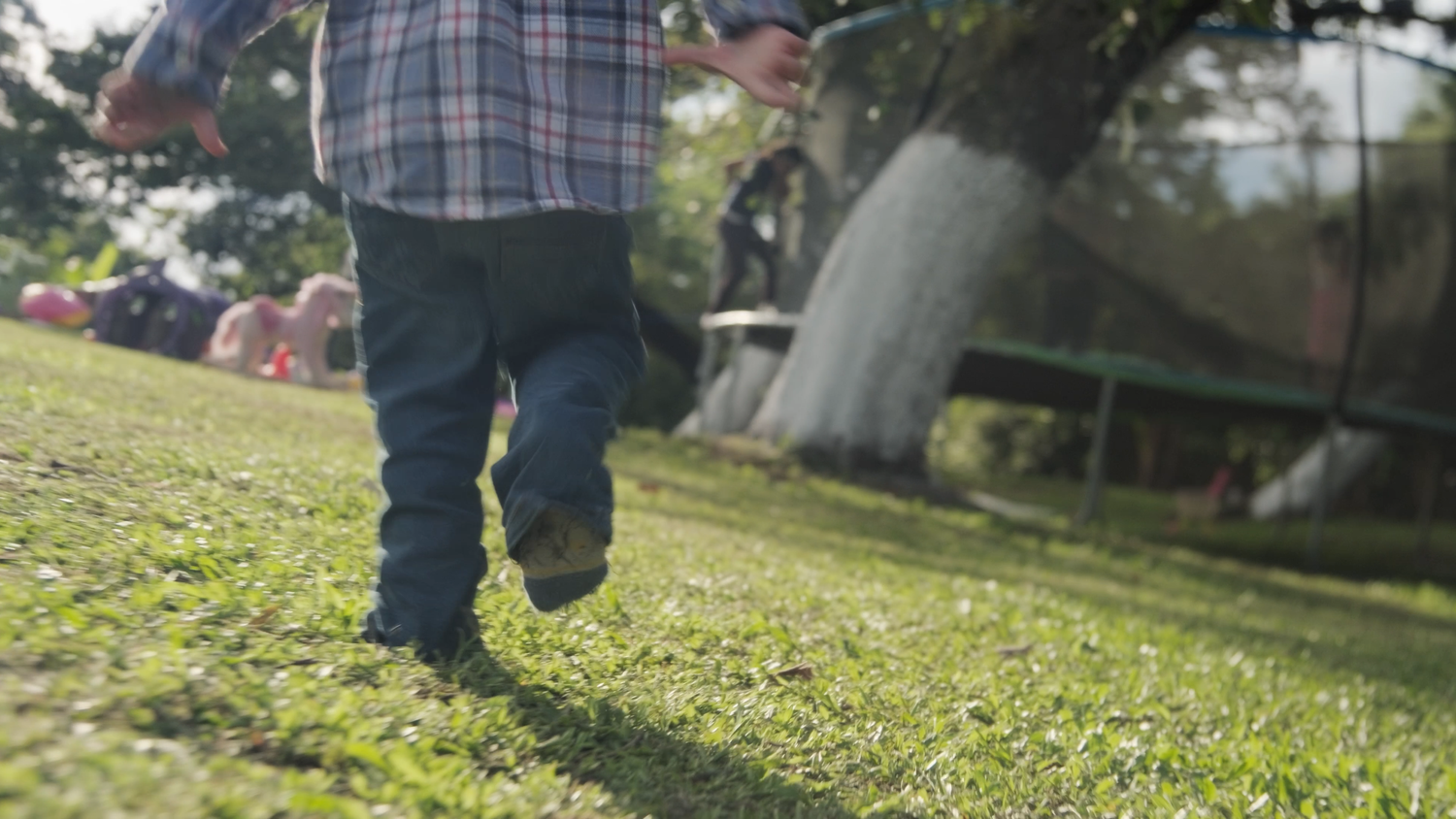 Toddler running on green grass in sunny backyard with trampoline in background.