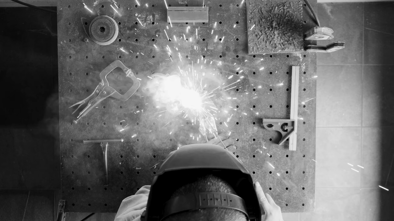Overhead black and white shot of a welder creating bright sparks at a metal workbench.