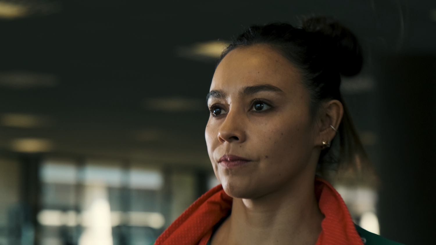 A focused female athlete with a bun hairstyle and orange towel gazes forward in a sports facility.