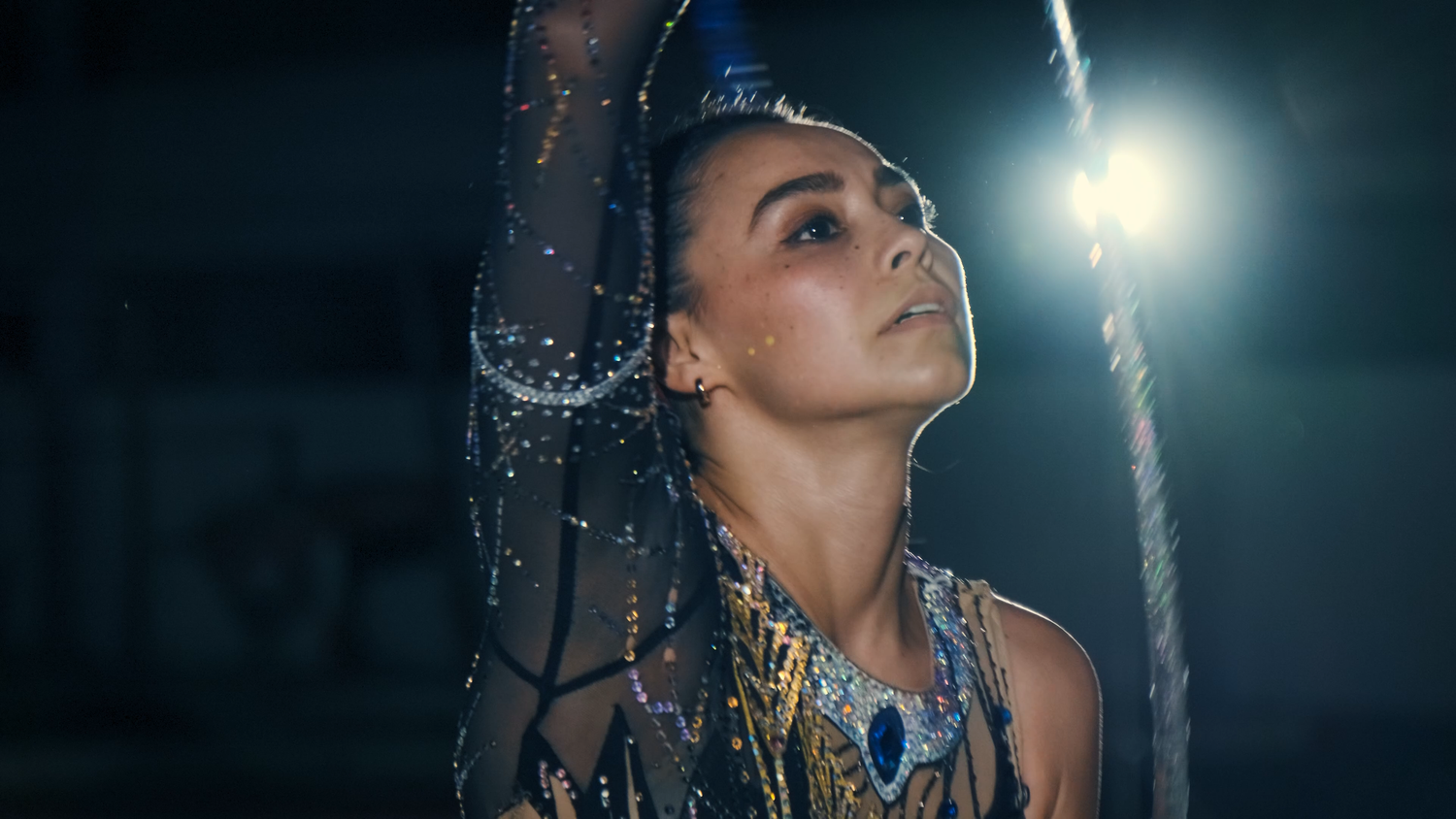 Focused female rhythmic gymnast in ornate blue leotard holds ribbon under dramatic spotlight.