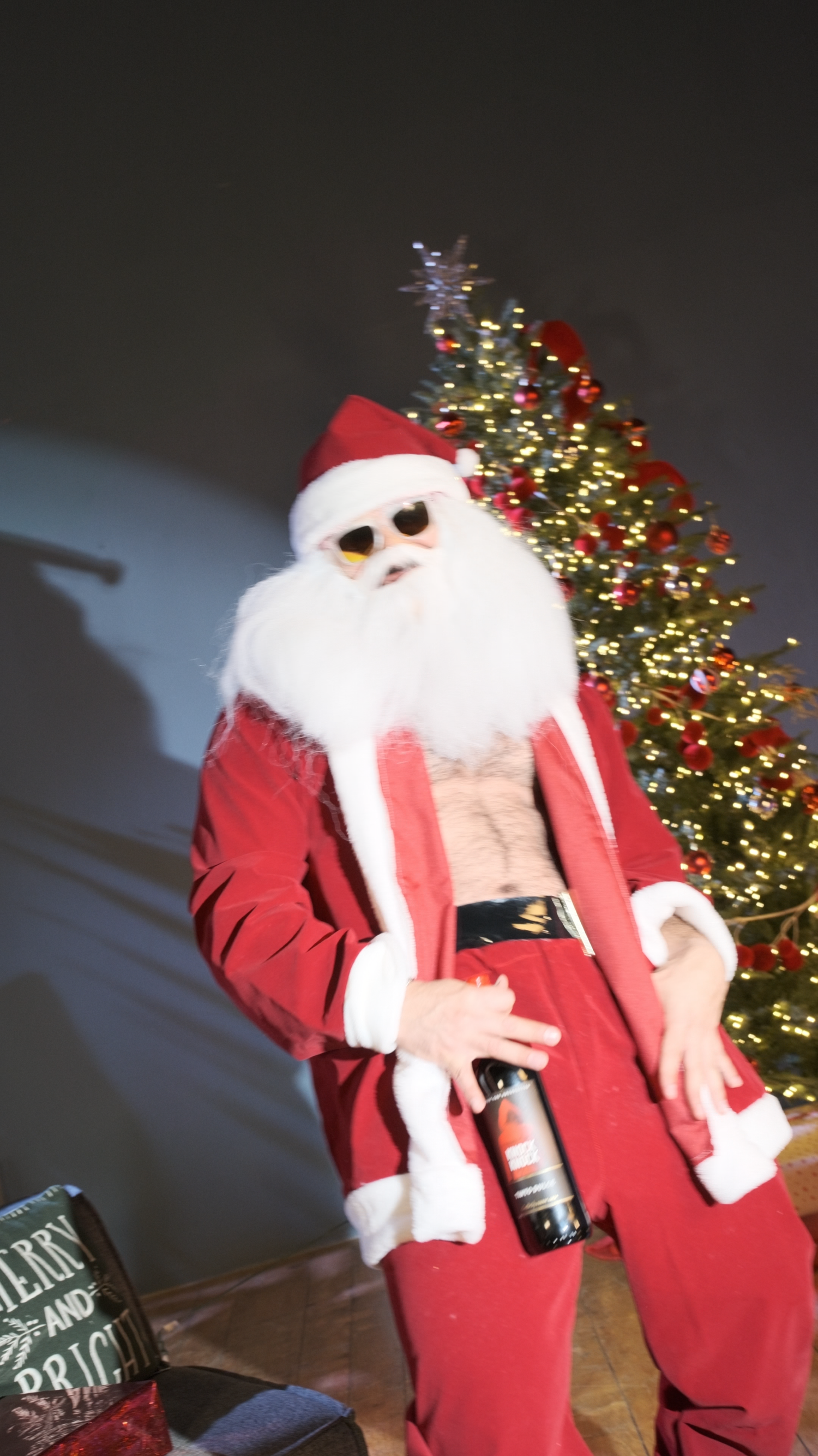 Person in open Santa costume with sunglasses holding a wine bottle in front of a lit Christmas tree.