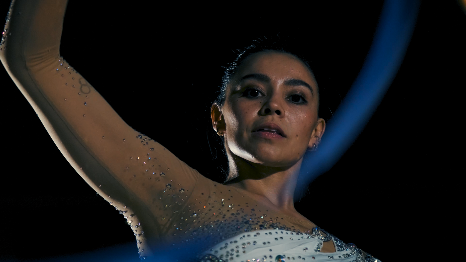 Elegant female rhythmic gymnast in sparkling white leotard performs gracefully under dramatic lighting.