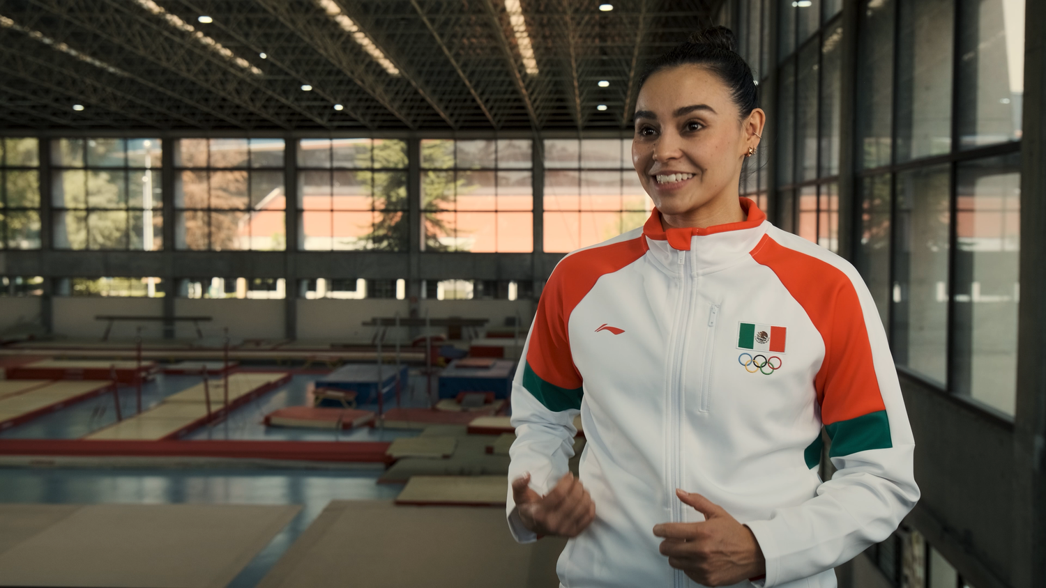 A smiling female athlete wearing a Mexican Olympic team jacket poses in a gymnastics training center.