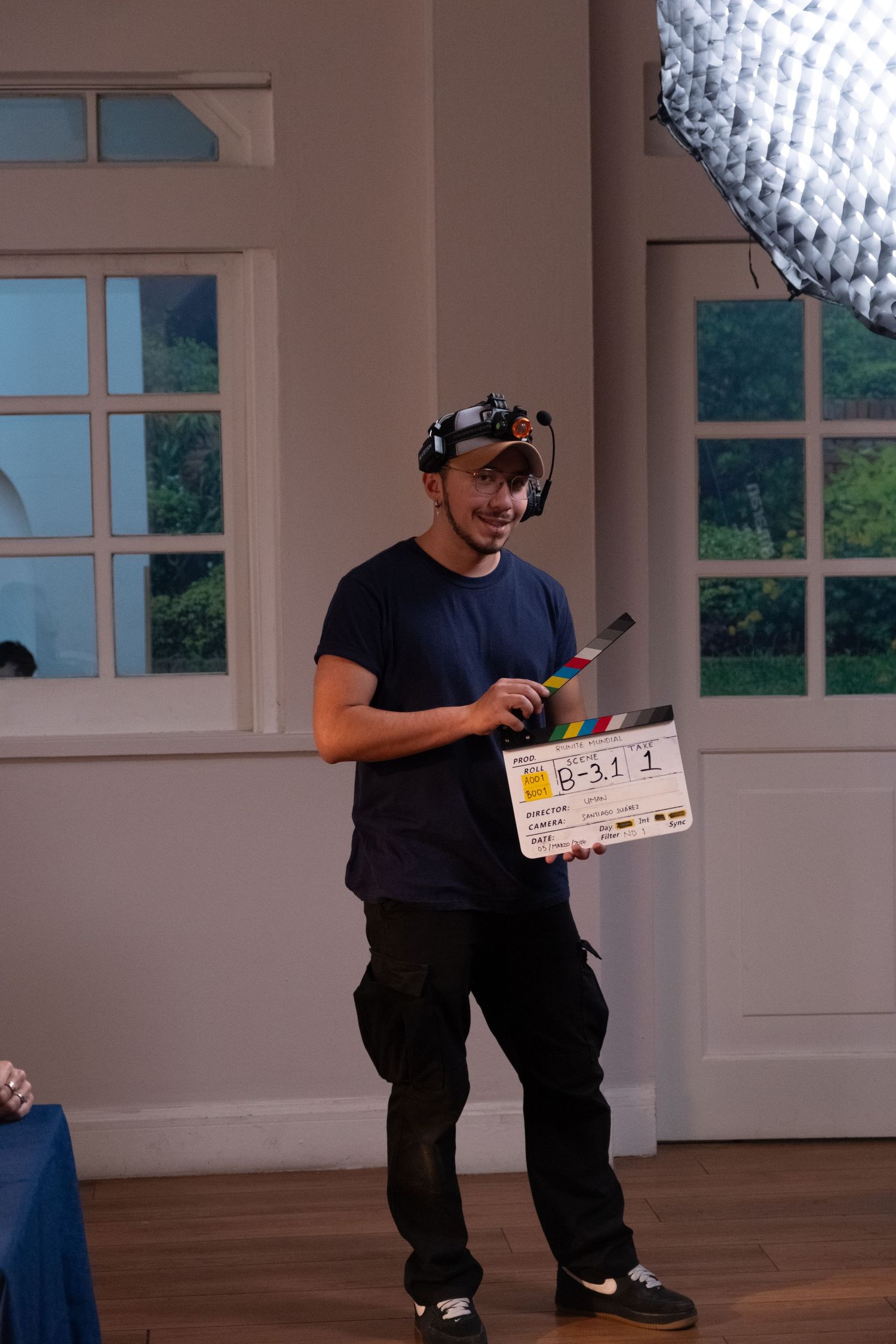 Film crew member holds a clapperboard on a bright indoor movie set with large windows.