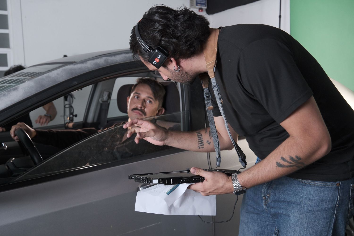 Film director reviewing a shot on a monitor with a mustachioed actor seated inside a car on set.