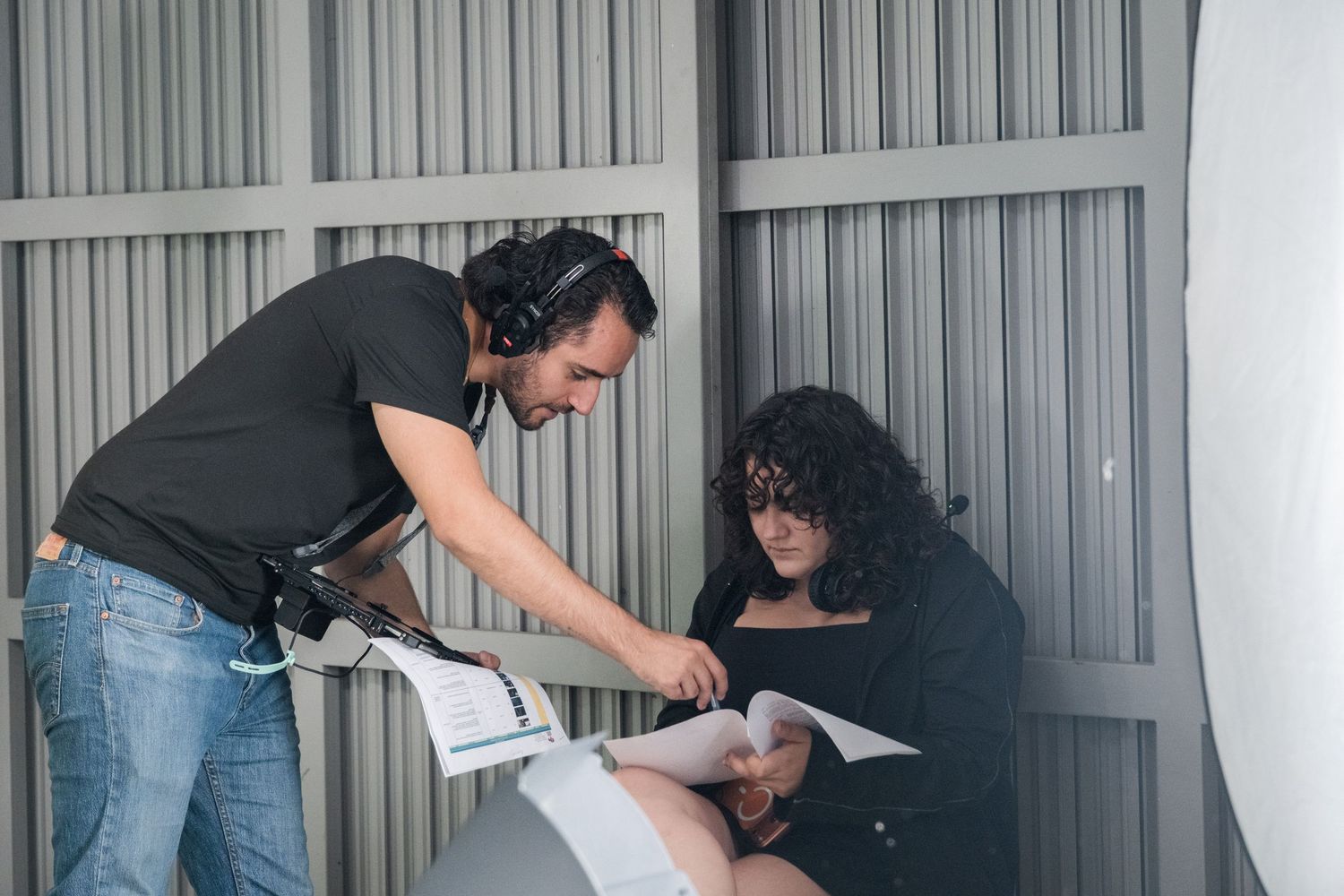 A film crew member wearing headphones reviews a script with a curly-haired woman on a film set.