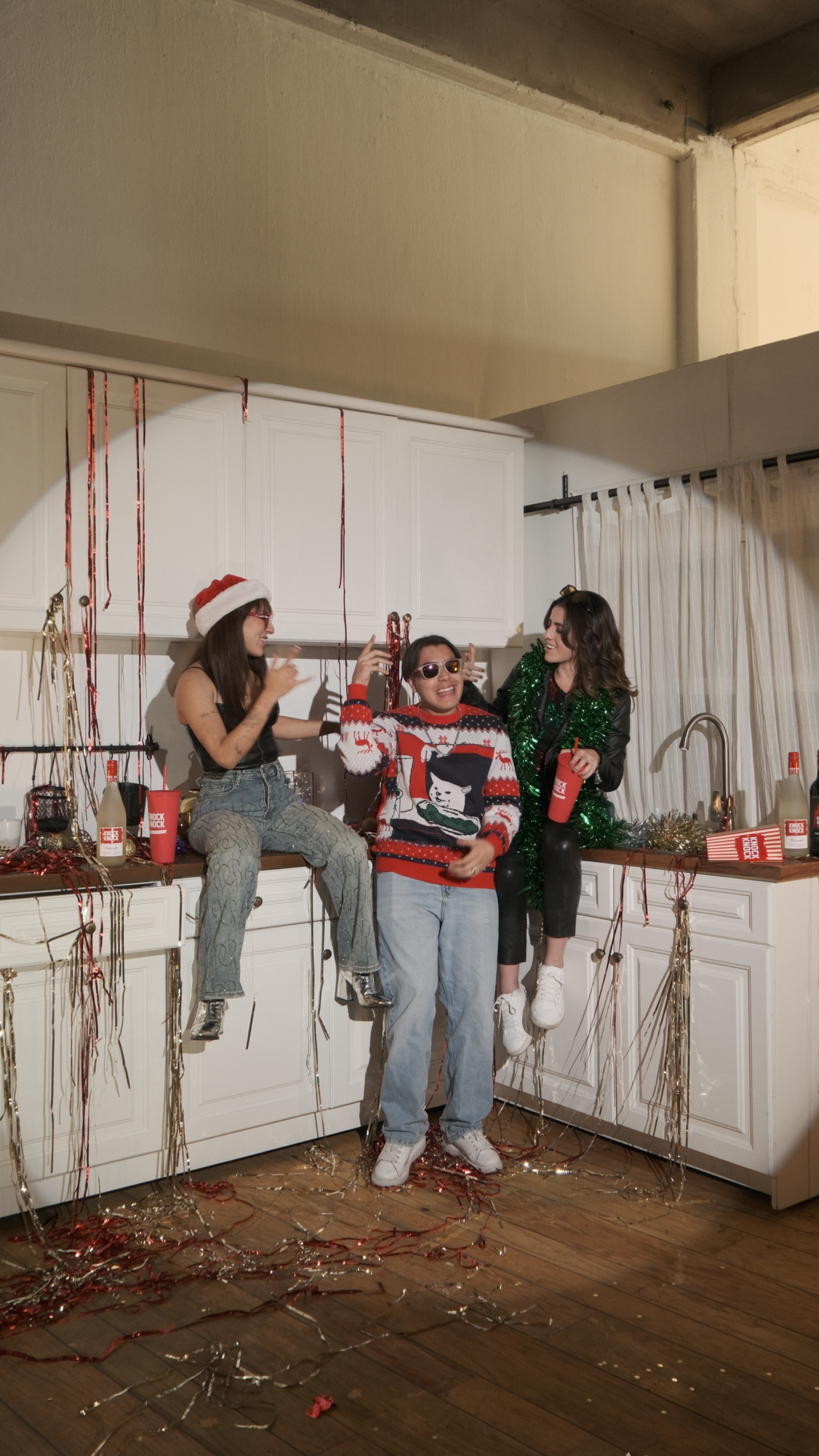 Three friends enjoying holiday drinks in festive kitchen decorated with Christmas streamers and ugly sweater.