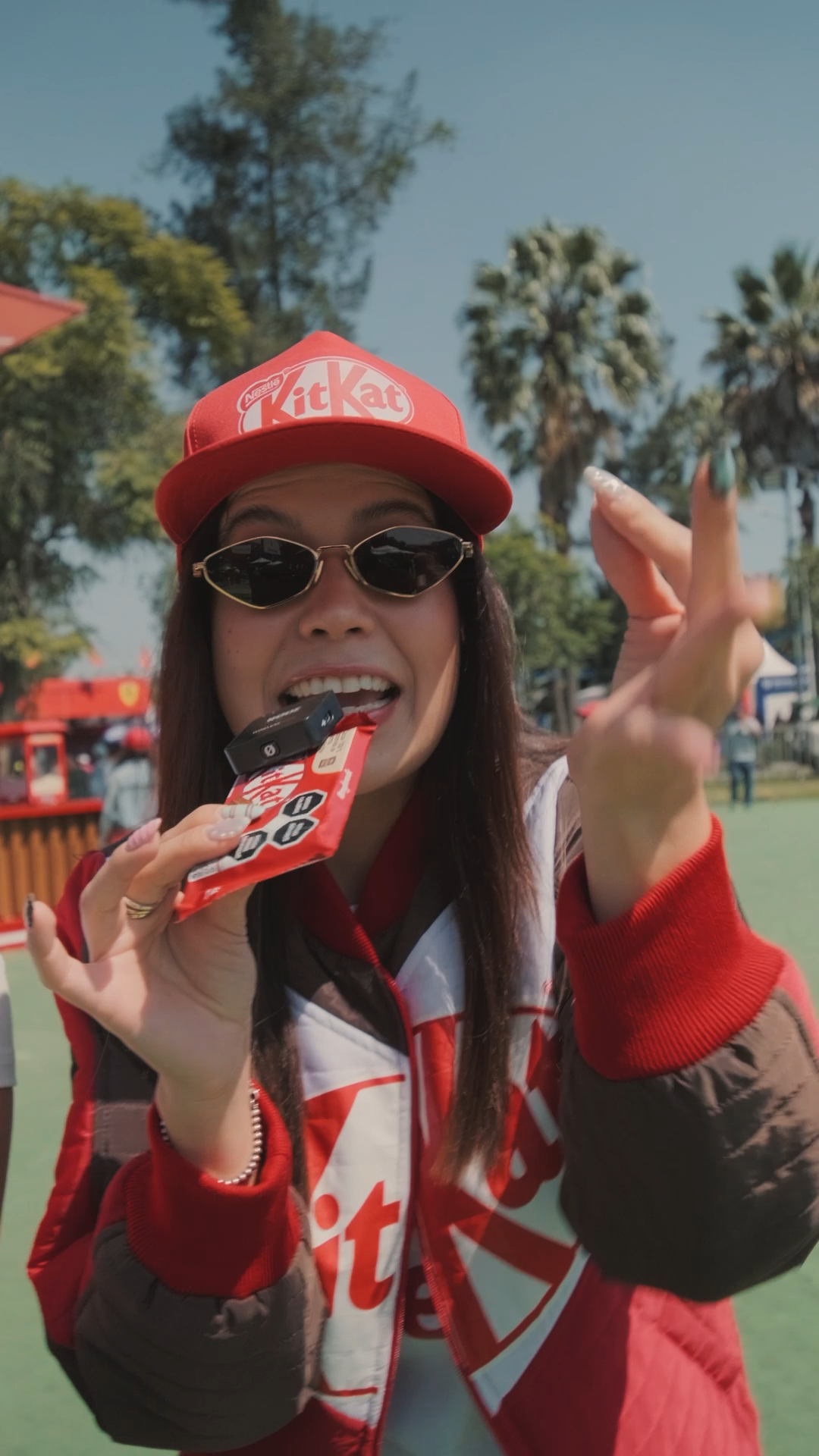 Woman in red KitKat branded cap and jacket eating a KitKat bar outdoors near palm trees.