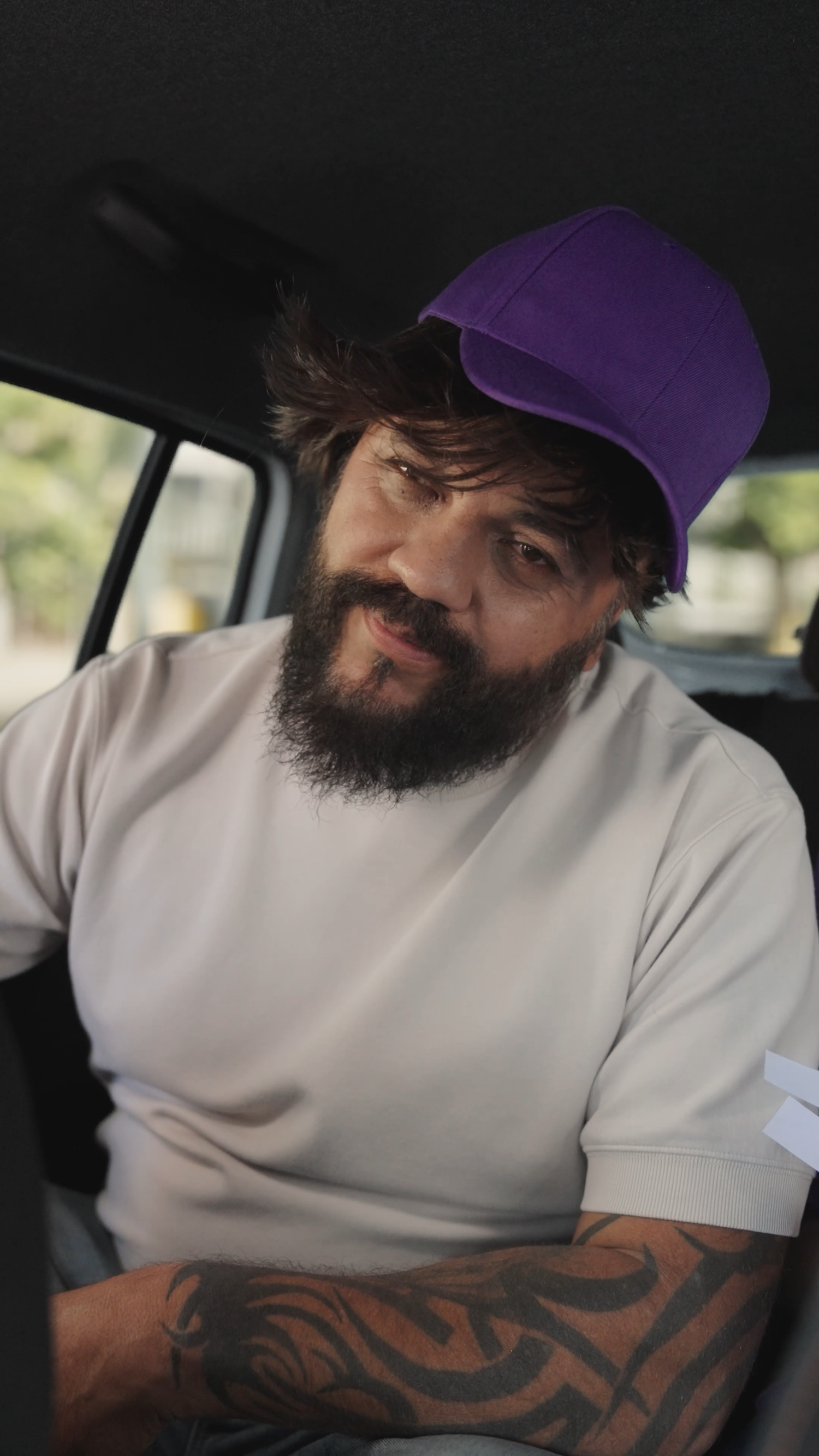 A bearded tattooed man wearing a purple cap smiles while sitting in the back seat of a car.