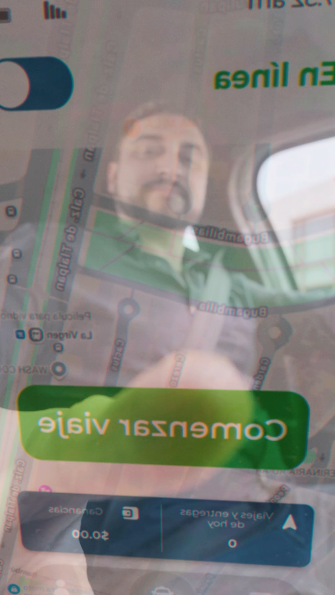 Reflection of a man in a car window showing a ride-sharing app with Comenzar viaje button.