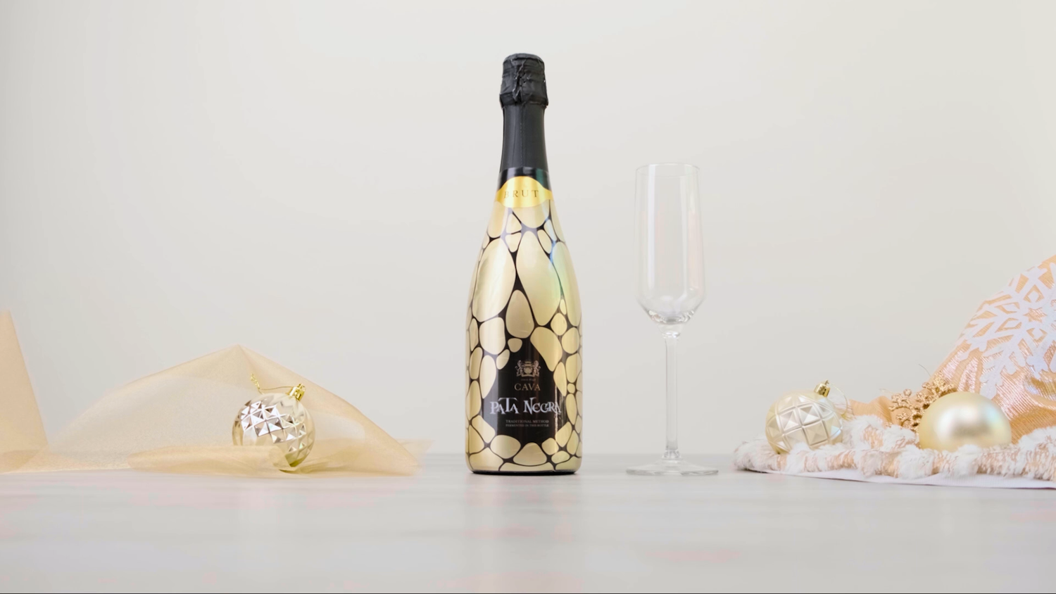 Gold geometric-patterned Cava sparkling wine bottle with champagne flute and holiday ornaments.