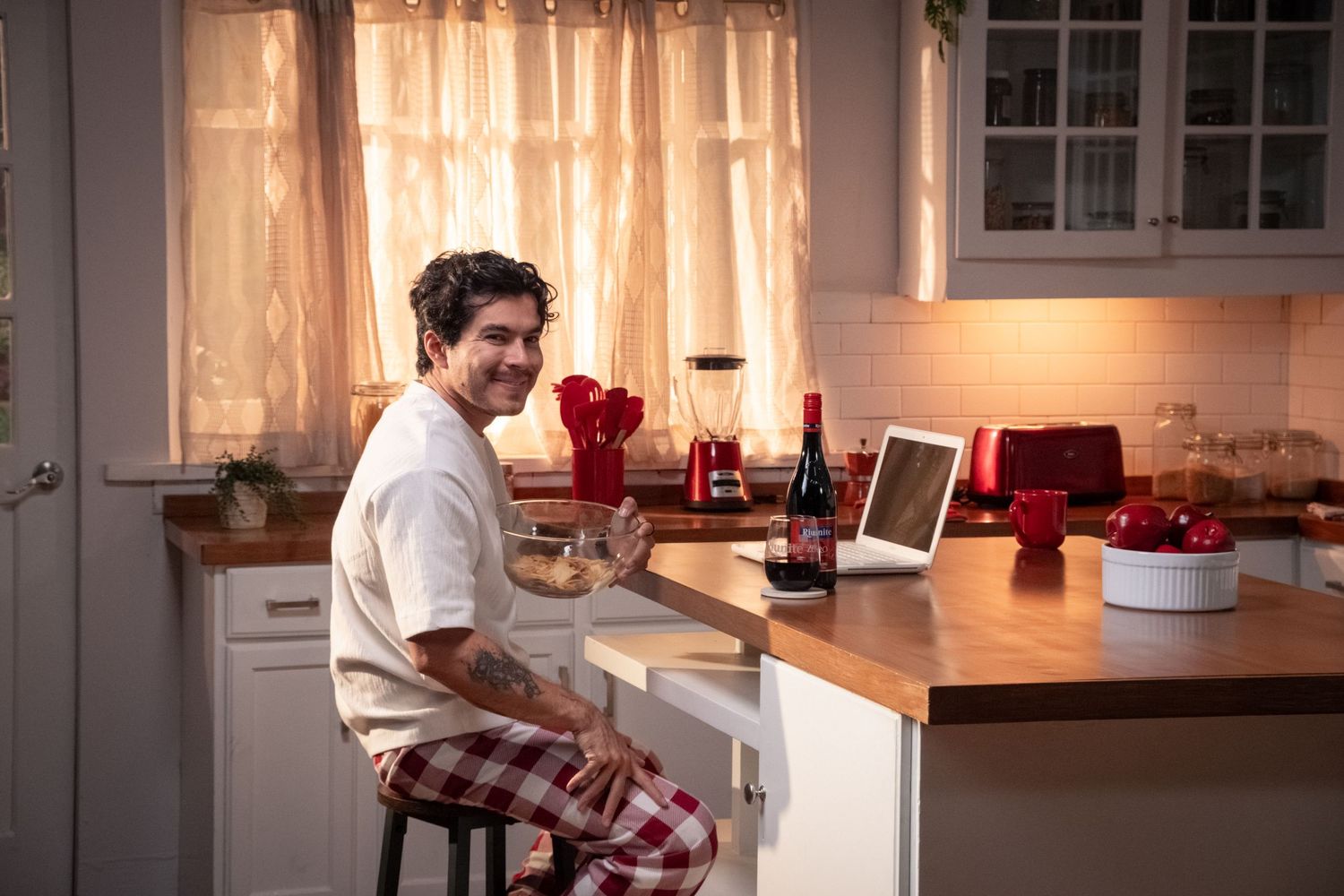Smiling man sits at kitchen counter with Riunite wine bottle and laptop.