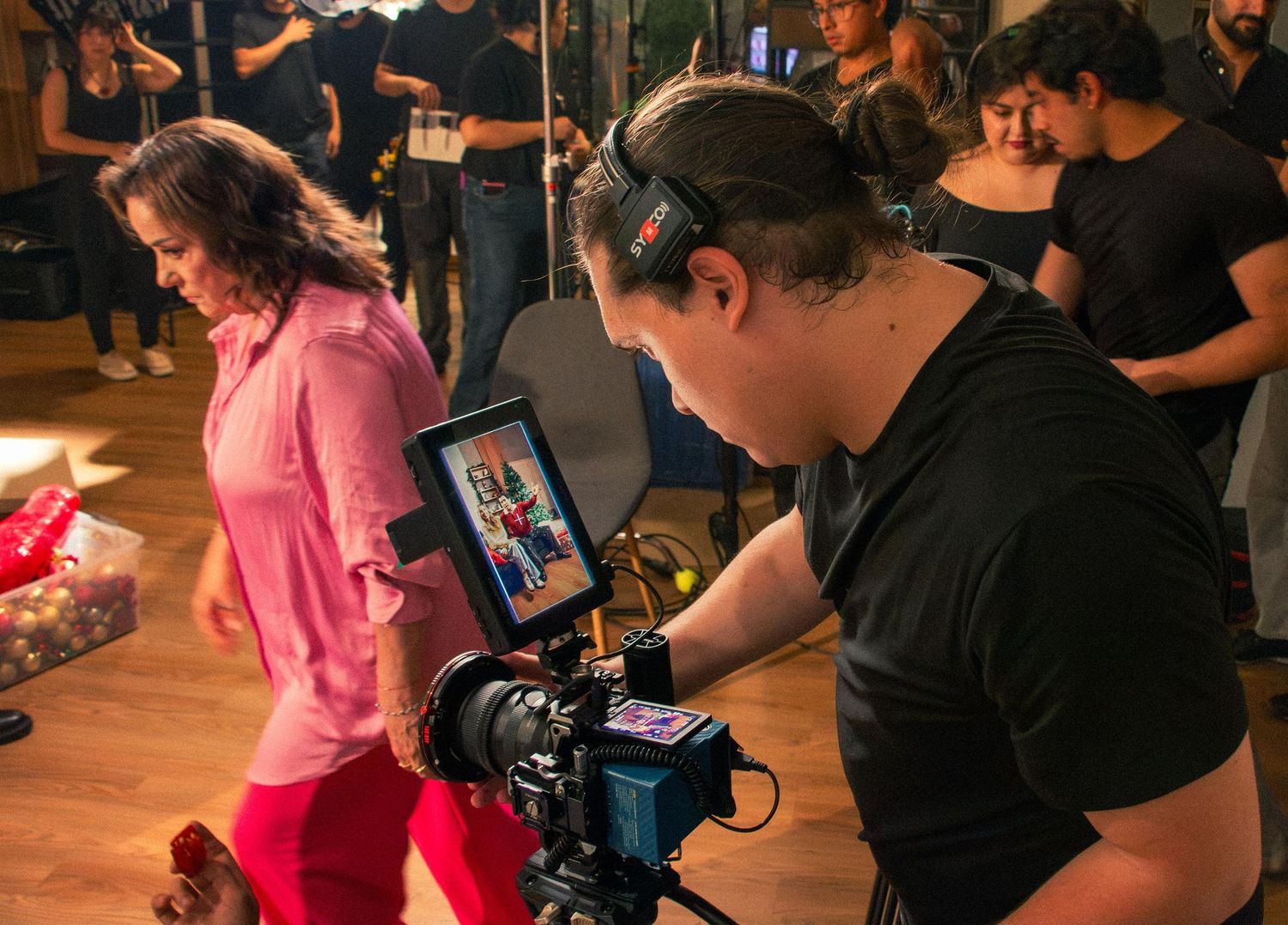 Behind-the-scenes photo of a cameraman filming a woman in pink on a vibrant indoor film set.