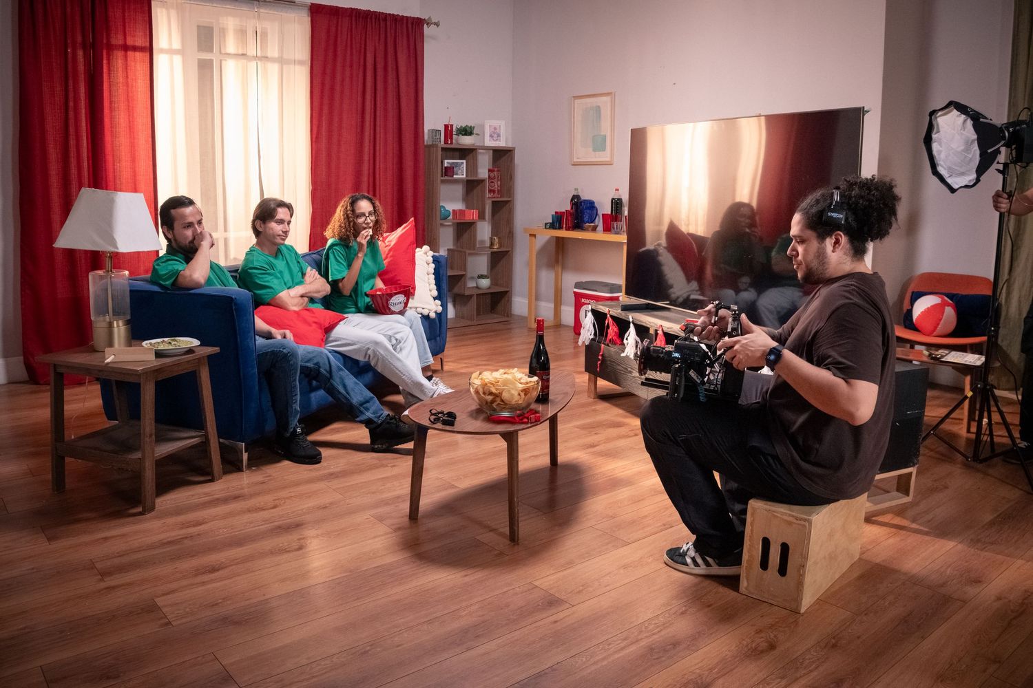 Cameraman films three actors on a living room set with red curtains and studio lights.